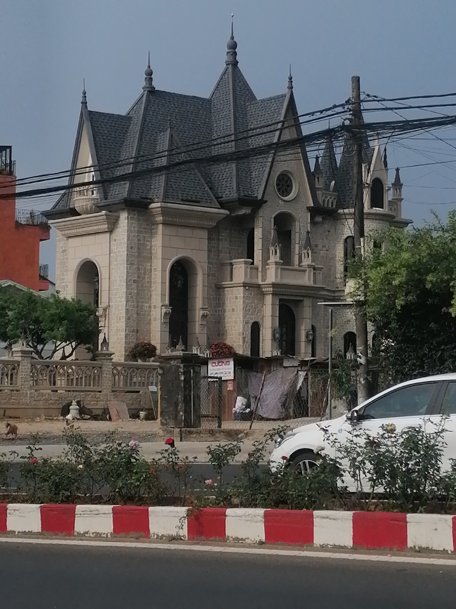 The image shows a gothic-style building in Da Lat, Vietnam. The building has a grey slate roof with multiple gables and spires. The exterior is constructed of light-colored stone blocks with arched windows and doorways. The building is surrounded by a low stone wall with a gothic-style railing. In front of the wall, there is a narrow garden bed with red and white painted curb. A white car is parked partially visible on the right side. Power lines run across the upper portion of the image. A small dog is seen near the wall. A sign with "Cuong" on it is visible near the entrance. An adjacent orange building is seen on the left. The sky is overcast.