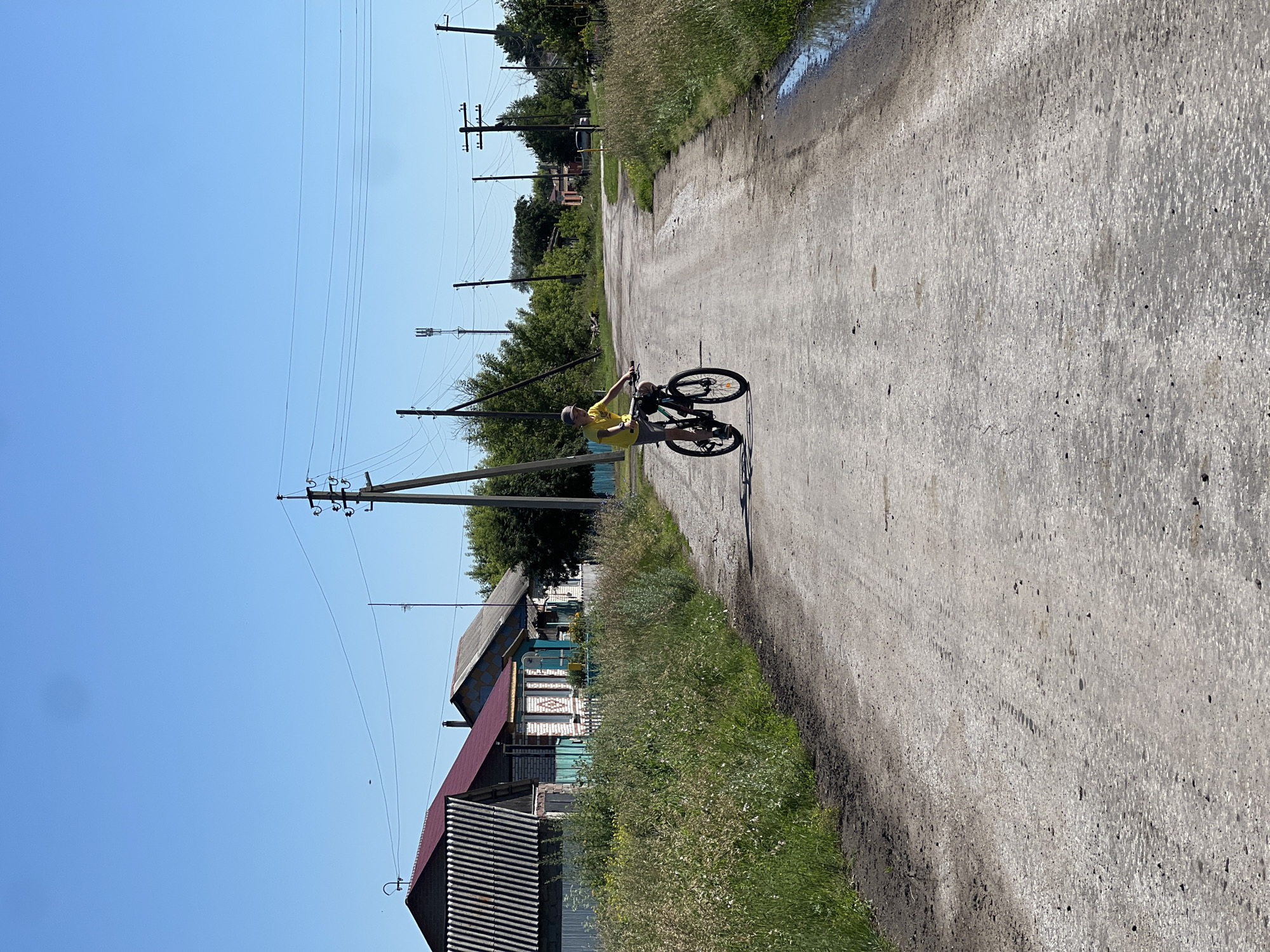 A young person in a yellow shirt and a grey cap rides a black bicycle down a dusty, rural road. The road is lined with tall, dry grass on both sides. In the background, several modest, colorful houses are visible, with traditional wooden fences. Telephone poles and power lines crisscross the clear blue sky. The overall scene suggests a quiet, sunny day in a village in Pivkino, Russia.