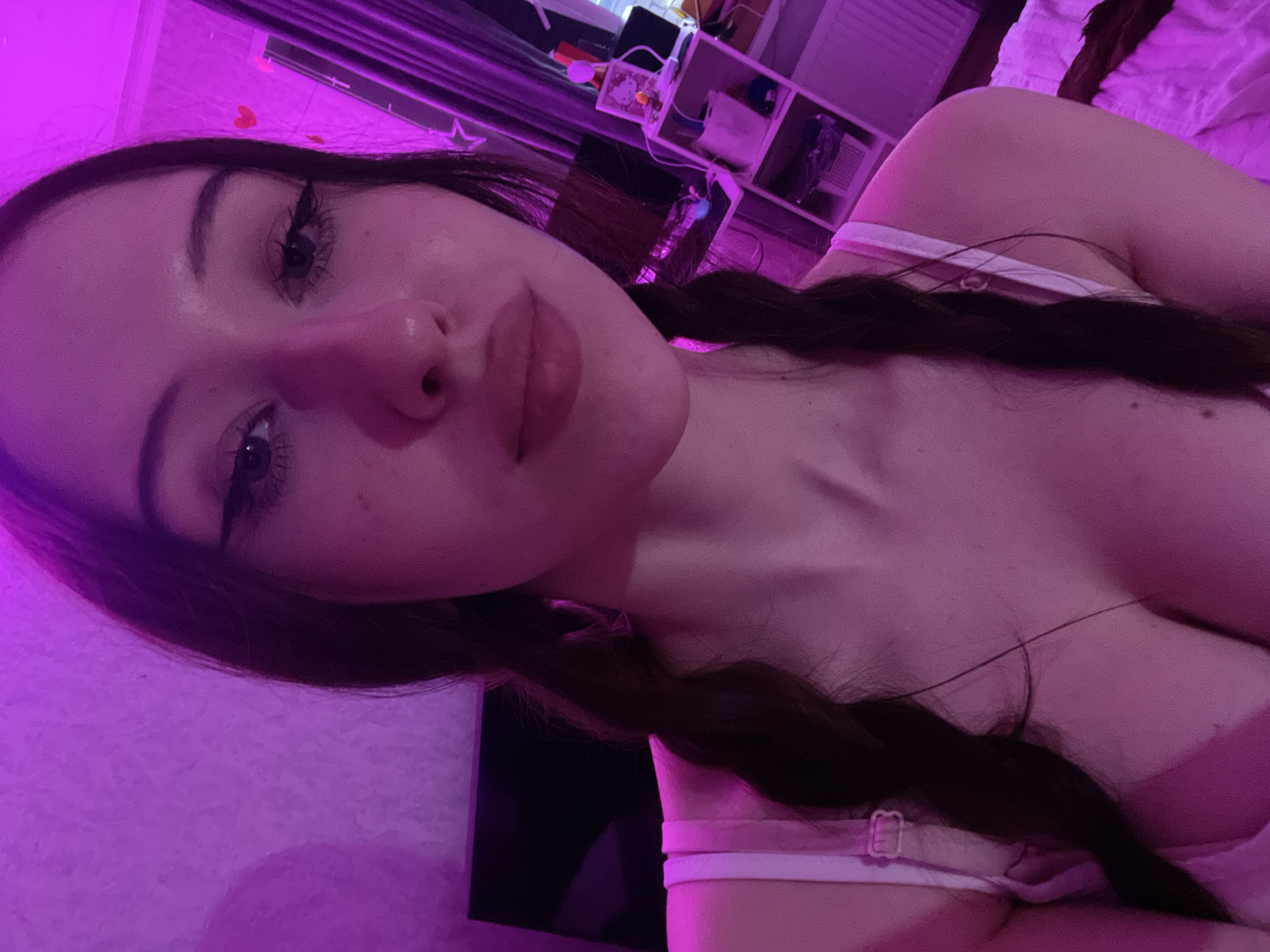 An indoor, close-up, chest-up portrait depicts a young female located in Votkinsk, Russia. The subject has dark hair styled into two braids, one visible over each shoulder, and fair skin, which is rendered with a prominent magenta hue due to the artificial lighting. She directly faces the camera, exhibiting distinct eyeliner, long eyelashes, and full lips. She is wearing a light-colored, possibly pink or white, two-strap garment, with a visible buckle on the left strap. The background is illuminated by a strong purple-pink light source. To the left, a textured wall with a dark, rectangular object (possibly a screen) partially visible. To the right, a window covered by vertical blinds, a white multi-tiered shelving unit displaying various items including a Hello Kitty framed picture, and a radiator are present. Small red heart cutouts are affixed to the wall near the window.