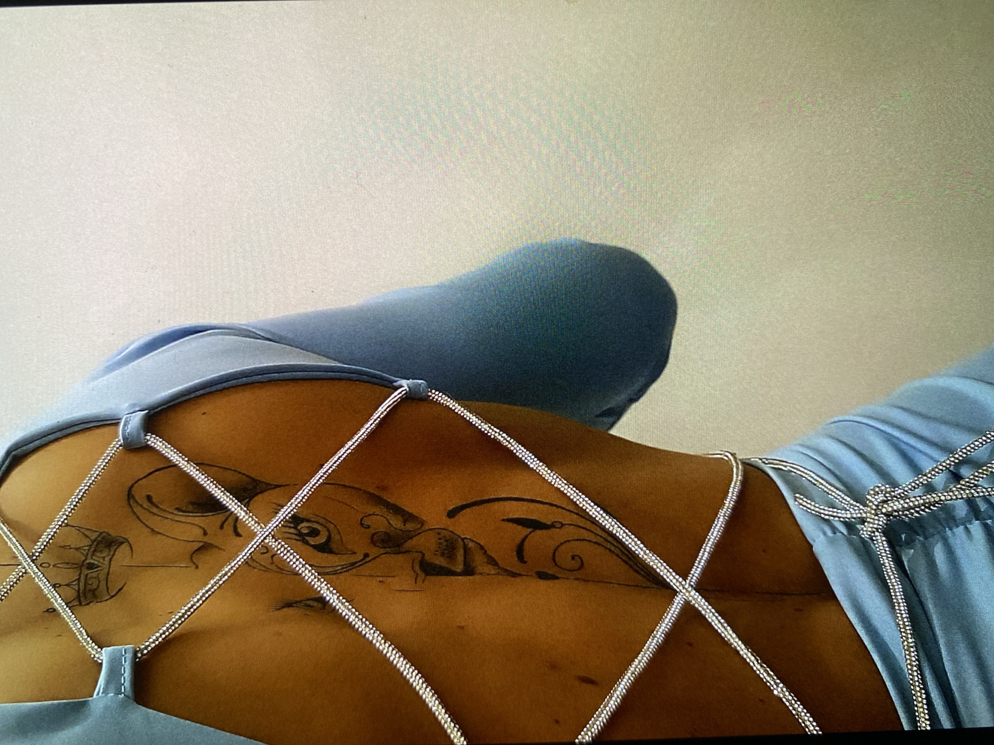 The image displays the lower back and shoulder area of a person, viewed from a slightly elevated perspective. The person is wearing a light blue garment, possibly a dress or top, that features intricate silver-colored, beaded straps crisscrossing diagonally across the back, forming a diamond pattern. On the right side of the frame, near the shoulder, the garment's fabric is gathered and secured by several tied silver straps.
A prominent black ink tattoo is visible on the person's lower back. The tattoo is horizontally oriented and depicts an ornate, stylized design. On the left side of the tattoo, there is a detailed element resembling an eye or a large oval shape, adjacent to what appears to be a crown or a similarly ornate structure. Moving towards the center and right, the tattoo incorporates flowing, organic lines, intricate swirls, and forms that suggest an insect or creature with segmented body parts.
The person has a medium to dark skin tone. The background is a plain, light-colored surface, indicating an indoor setting. The lighting is bright and even. The image quality suggests it might be a photograph of a digital screen due to the visible pixel grid texture. There is no visible text.
