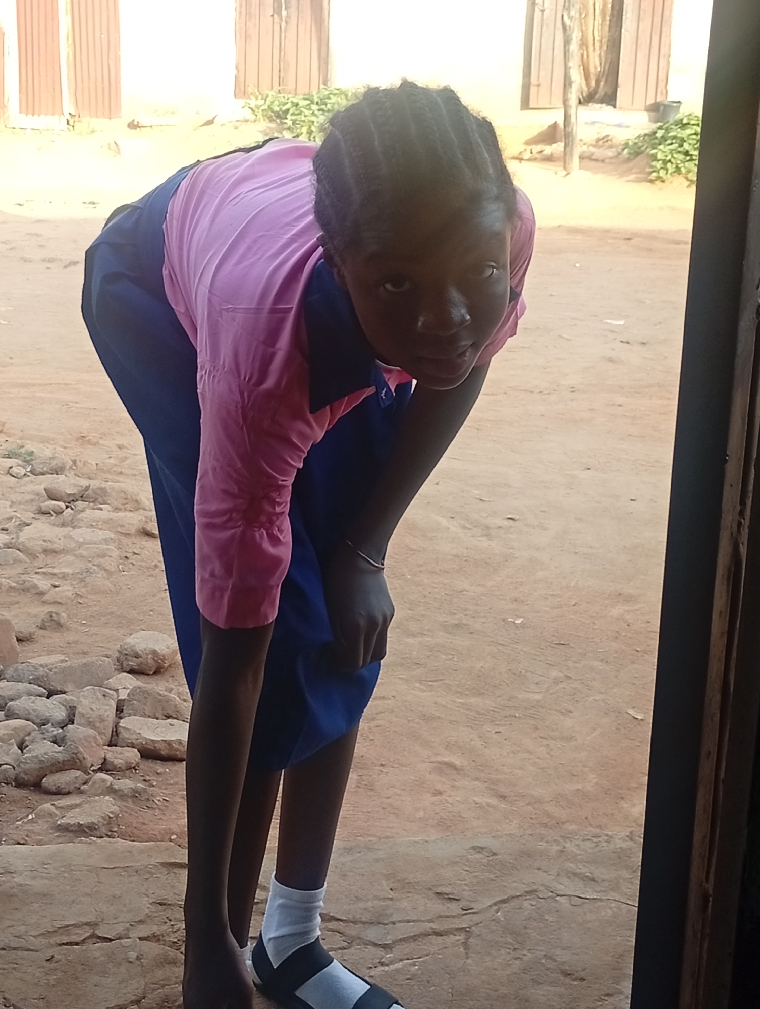 A young woman bends forward while standing near a doorway on a dirt ground in Unknown, Unknown. She is wearing a pink long-sleeved shirt with a dark blue collar, a dark blue skirt, and a white sock with a black sandal on one foot. Her hair is braided. To the left of the woman, there are several rocks. In the background, there are buildings with brown wooden doors and windows. Green bushes are visible behind the buildings.