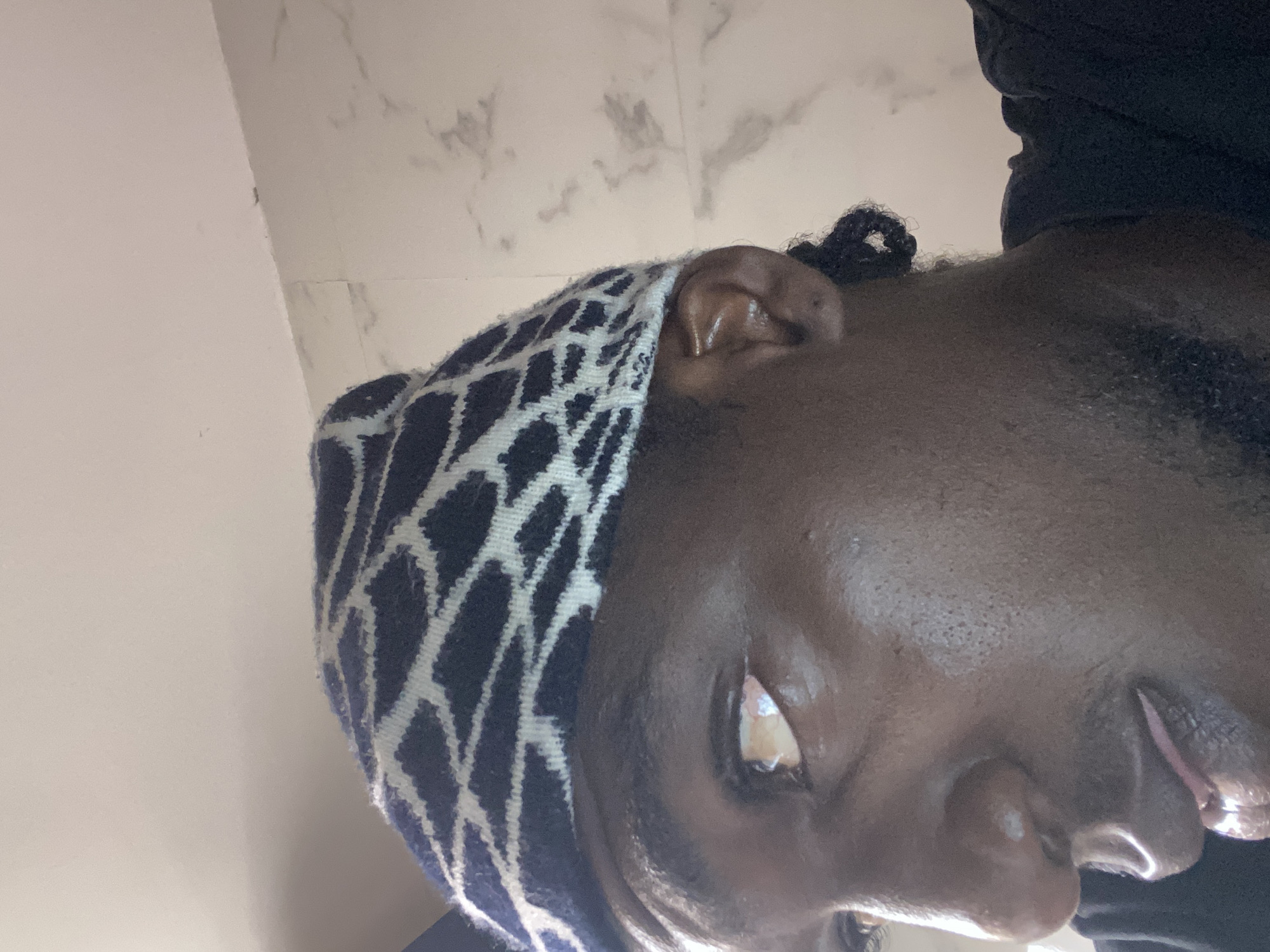 A close-up, upward-tilted shot of a person's face. The person is wearing a patterned head covering in dark and light colors. A section of their face is visible, including one eye, the nose, and part of the lips. The person appears to have dark skin. The background consists of a wall with a mottled texture. No specific location in Lagos, Nigeria can be determined from the image.