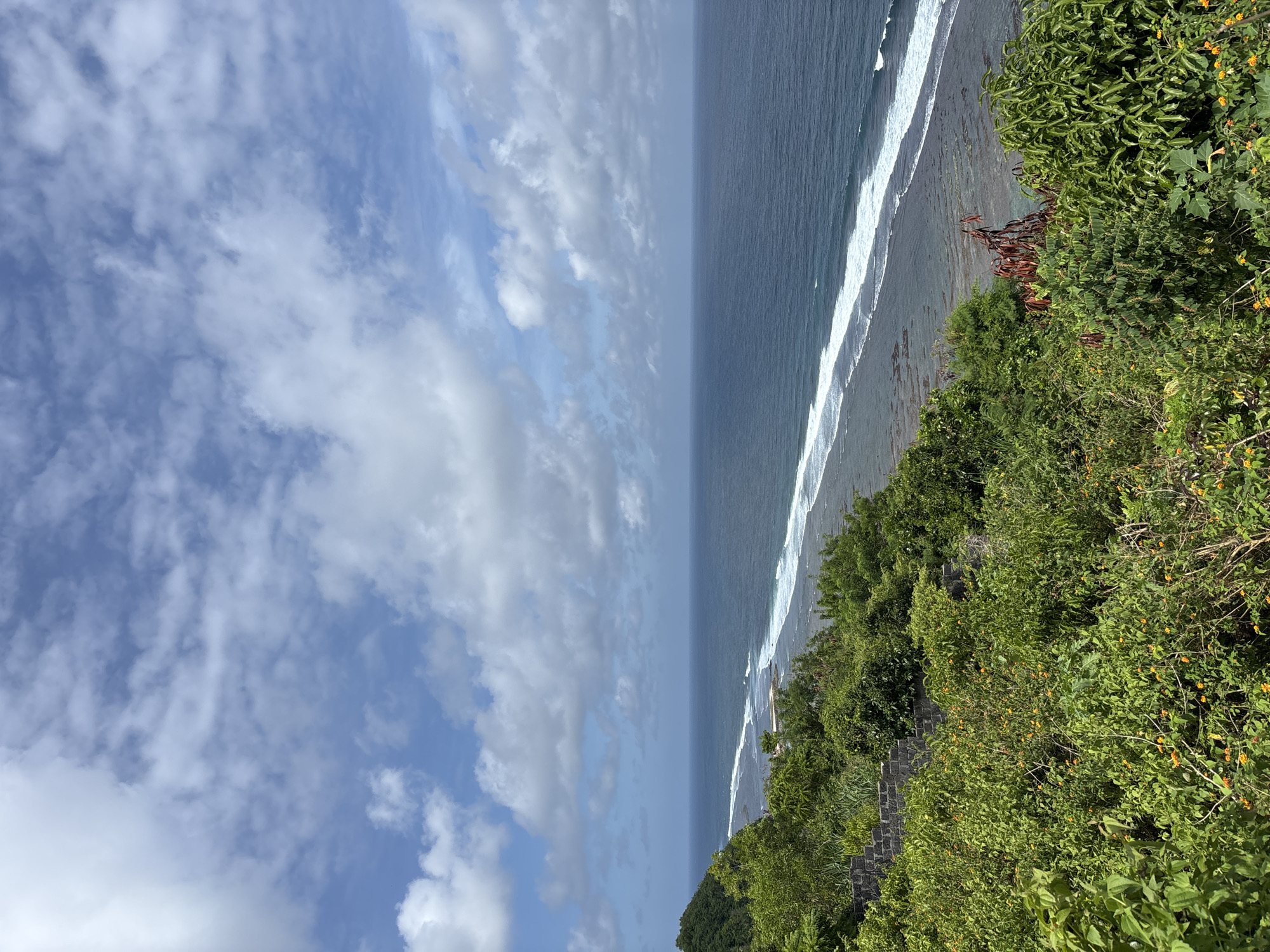 A high-angle, medium shot depicts a coastline under a partially cloudy sky. To the left, the sky is a mix of blue and white clouds. The ocean stretches from the center towards the upper-right, with visible waves breaking near the shoreline. Lush, green vegetation covers the area on the right side of the frame. A stone structure is also visible amid the greenery. The location is Kuta Selatan, Indonesia.