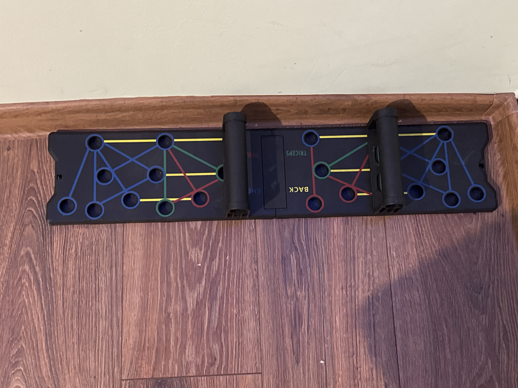 The image captures a black, multi-functional push-up exercise board resting on a wooden floor, positioned against a light brown wooden baseboard and an off-white wall. The board is rectangular and features a grid of circular holes and interconnected lines, color-coded to indicate target muscle groups for exercises.
Two black push-up handles are inserted into the board, ready for use. Visible text on the board, oriented upside down from the viewer's perspective, labels the different sections: "TRICEPS" is clearly legible, "BACK" is also visible, and partial text like "SHOUL" (for Shoulders) and "CHE" (for Chest) can be discerned. Blue lines and holes are associated with shoulders, red with chest, green with back, and yellow with triceps. The handles are currently placed within the areas corresponding to the triceps and back exercises.
The wooden floor exhibits a distinct grain pattern, suggesting a natural wood or laminate finish. The lighting in the room is ambient, indicating an indoor setting, likely a home gym or general living space. No people are present, and no activity is taking place; the equipment is stationary. The location context provided is Yana, Bulgaria, although no specific visual elements within the image itself confirm this geographical detail.