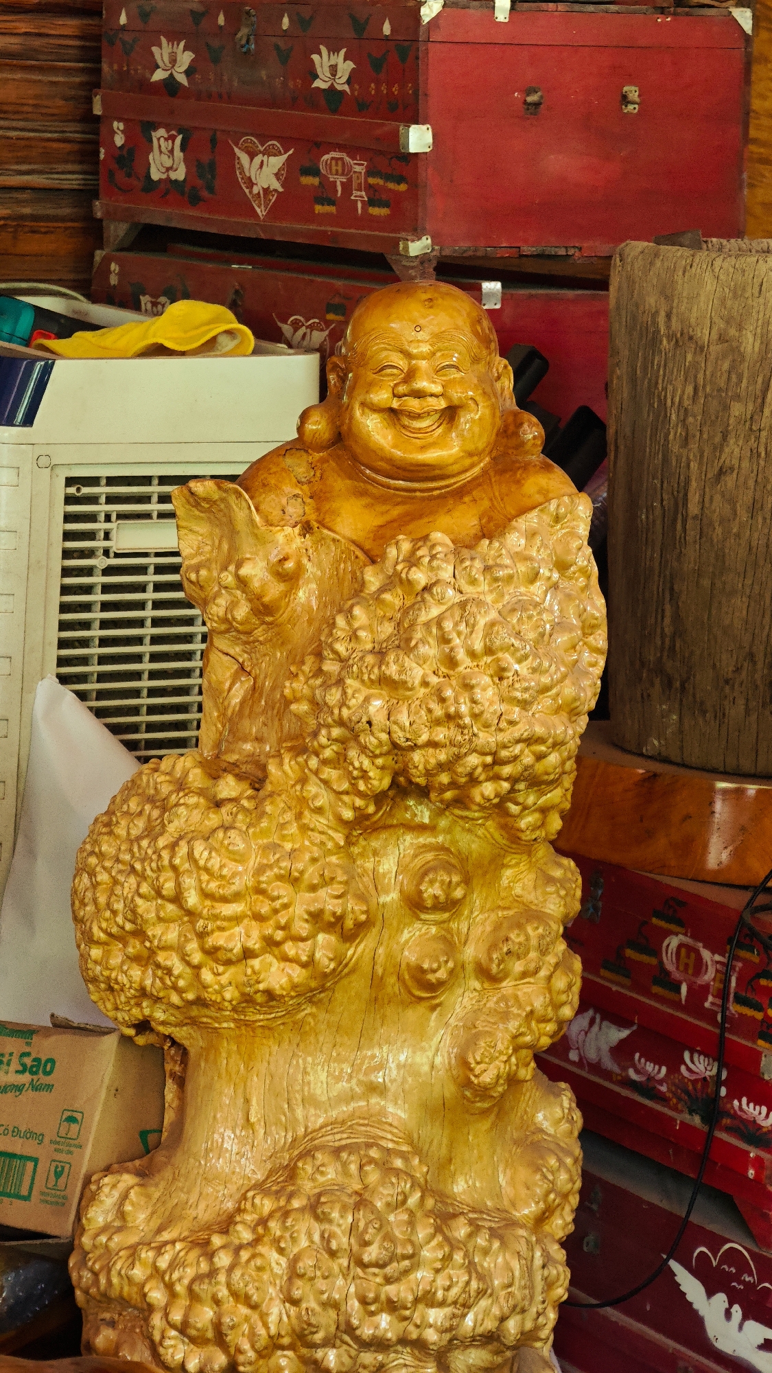 The image captures a detailed, full-length view of a large, golden-toned wooden statue of a smiling Hotei (Laughing Buddha) located in Bản Đêu, Vietnam. The Buddha statue is carved from what appears to be a natural wood burl, featuring a wide, jovial smile with visible teeth, prominent earlobes, and a rotund figure. The lower body and base of the statue are characterized by highly textured, knobbled, and organic wood formations, all coated with a glossy, golden-hued varnish.
In the immediate background to the left of the statue, a white rectangular air conditioner unit with a visible vent grille is present. Above it, a yellow cloth is draped over an unseen object, and parts of blue and white items are also visible. A brown cardboard box with Vietnamese text and a recycling symbol is partially visible at the bottom left, behind the statue's base.
To the right and in the upper background, several ornate red wooden chests or trunks are stacked. These chests are adorned with hand-painted motifs in white, green, and black, depicting lotus flowers, geometric patterns, stylized hearts, and an emblem resembling a lantern with an 'H' inside. A rough-hewn, cylindrical wooden post stands vertically to the right of the statue, exhibiting distinct wood grain. A black electrical wire runs diagonally across the right side of the frame, partially obscuring the red chests. The scene is brightly lit, revealing an interior setting with a combination of decorative and utilitarian objects.