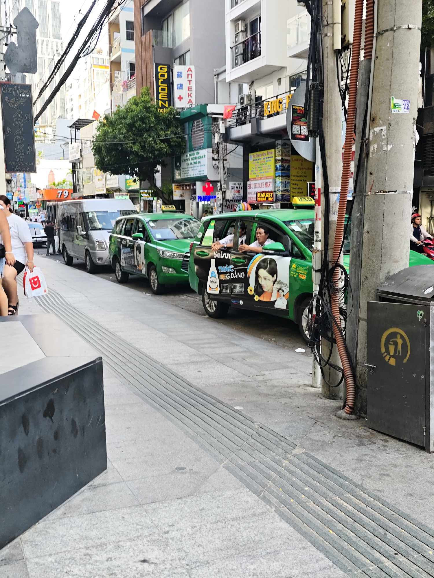 In Nha Trang Ward, Vietnam, on a street with buildings on the right and a sidewalk on the left, a line of green taxis is parked. The closest taxi has advertisements for "Sotur" and features images of women. A person in a white shirt is seated in the driver's seat, and another person, likely a passenger, is visible. Further down the street, a silver minivan is parked. A person in a white t-shirt and dark shorts is walking on the sidewalk, carrying a red and white shopping bag. The street is paved with concrete and features a tactile paving strip for the visually impaired. Electrical wires are suspended overhead, and a concrete utility pole with a trash receptacle is situated on the sidewalk. The general environment suggests a commercial area within a city.