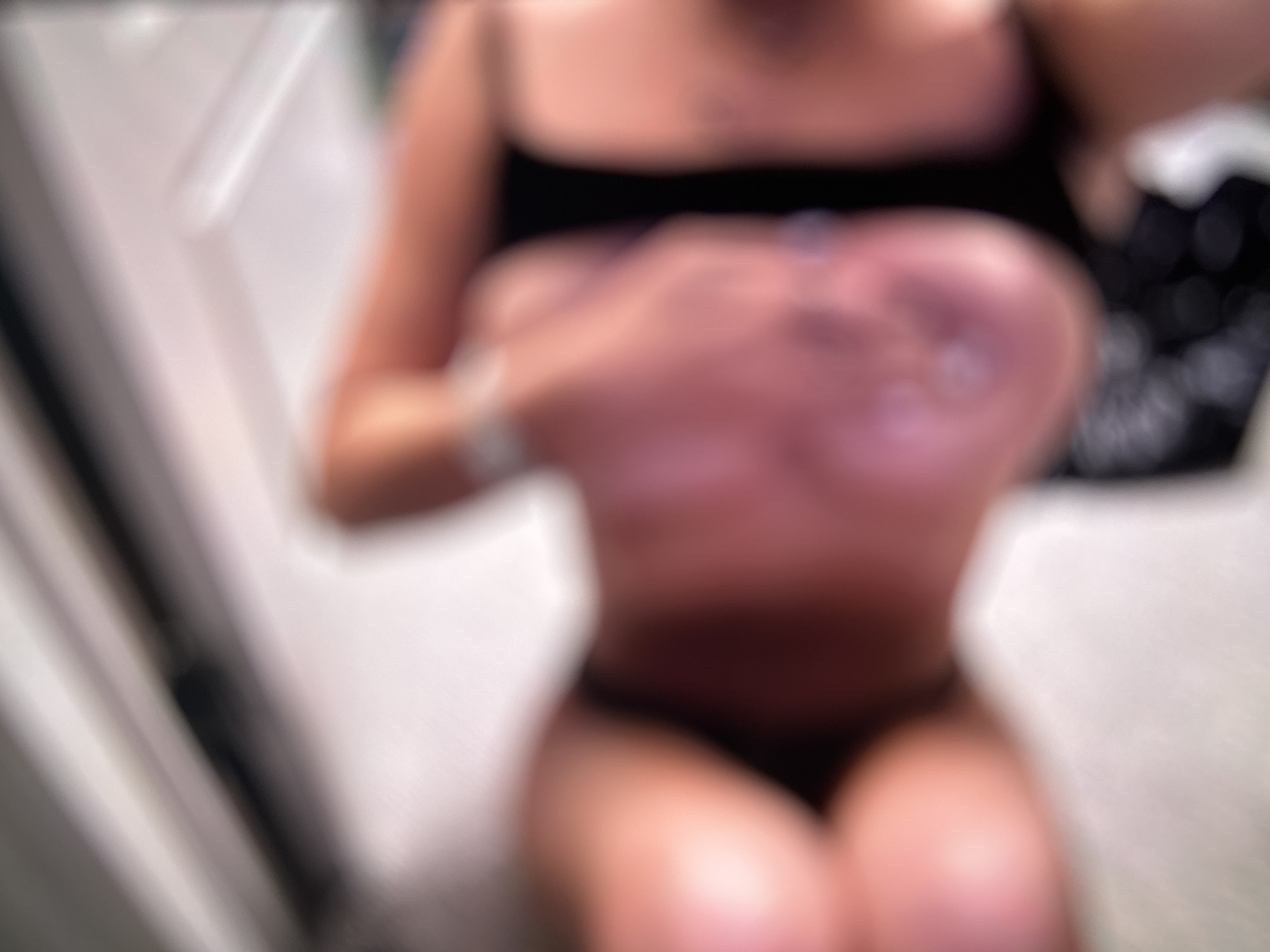 The image depicts a severely blurred, close-up scene indoors in the city of Blagoevgrad, Bulgaria. The primary subject appears to be a person, rendered in soft, pinkish-peach skin tones. The image is oriented such that a partially visible, inverted face, identifiable by discernible lips and the area of the nose, is centrally located. A hand with a silver-colored ring on one finger is visible near the top-center of the frame. To the right, a dark strap, possibly part of clothing like a bra or tank top, is visible against a blurred skin-toned area. The background elements include light, possibly white, architectural features and some dark areas with circular bokeh, suggesting diffused light sources. The extreme lack of focus prevents identification of specific actions or detailed environmental context beyond an indoor setting.