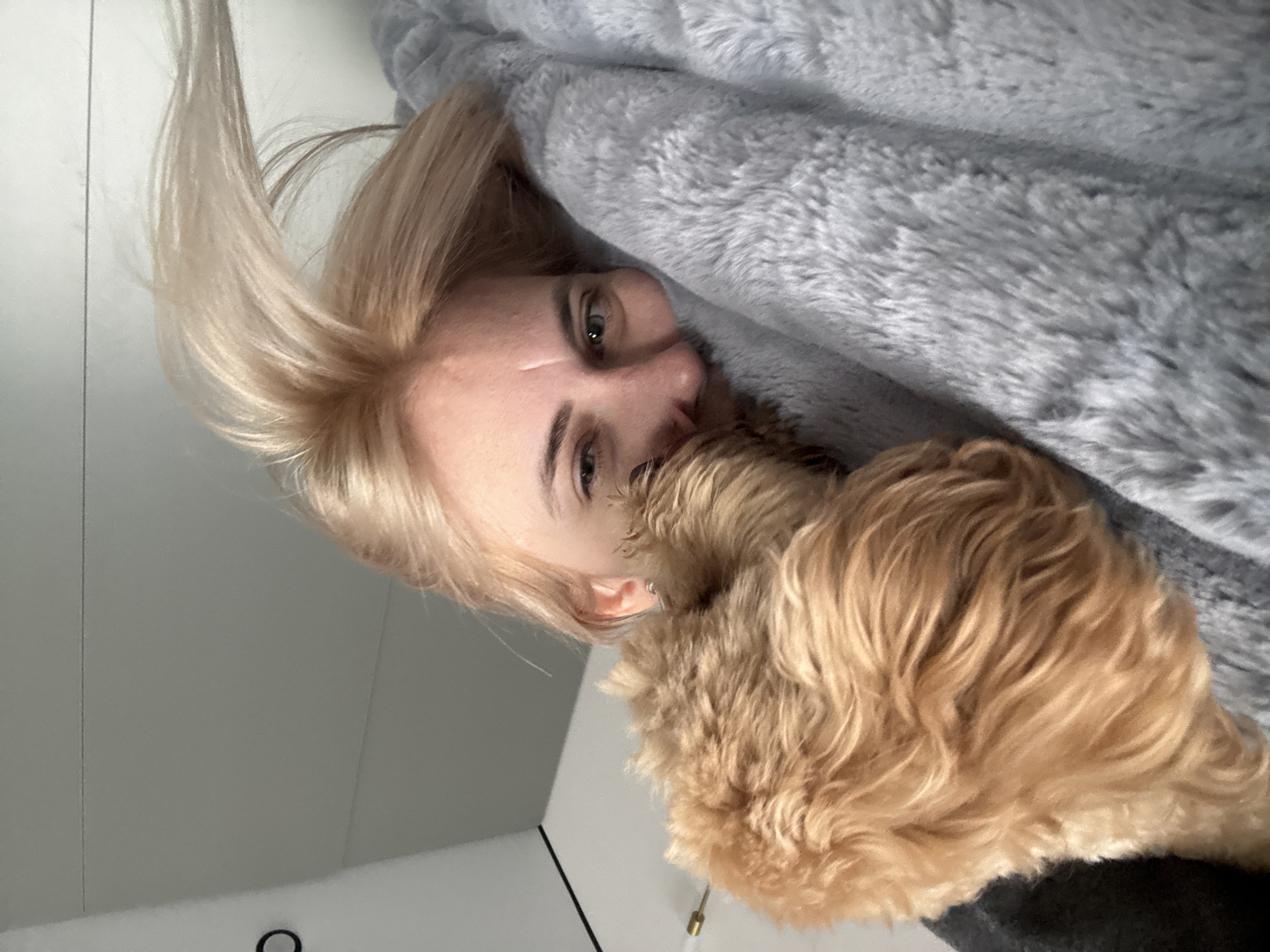 A woman with blonde hair lies down next to a light brown dog. The woman is partially covered by a gray, fluffy blanket. The background includes a white wall and a door frame with a light fixture. No information is available to determine if this image was taken in Saratov, Russia.