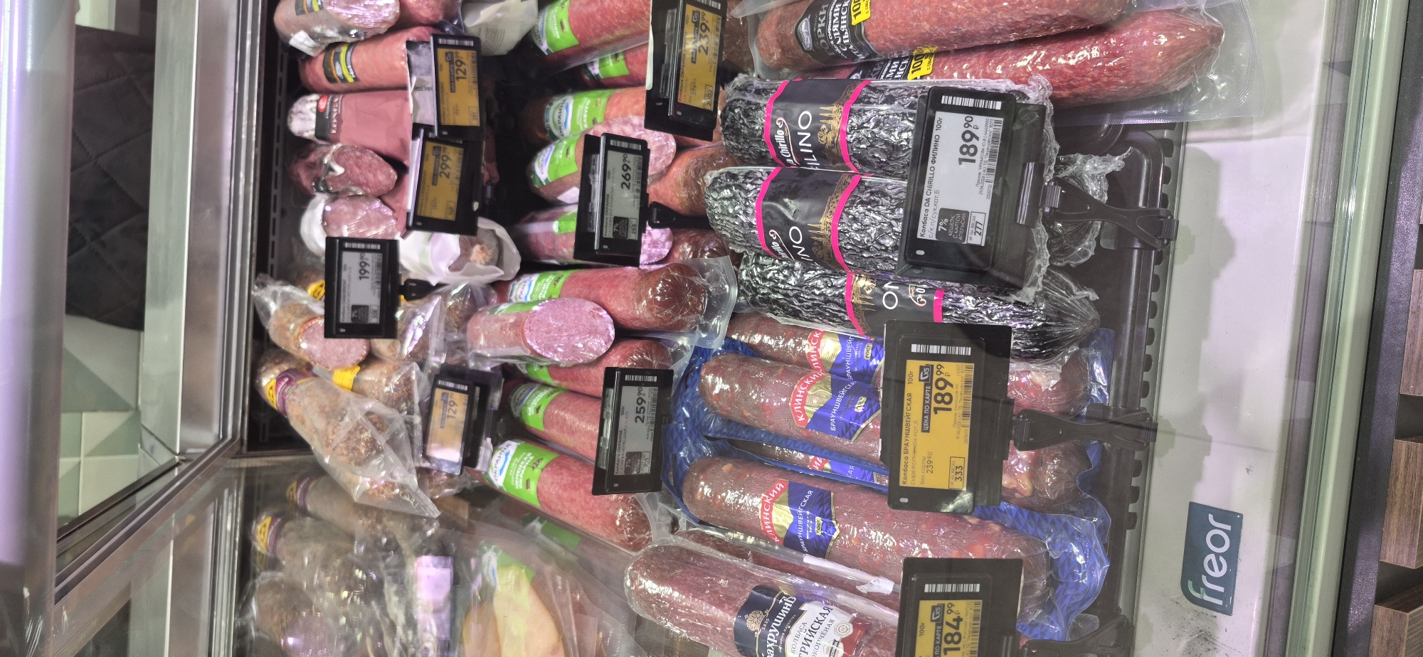 This is a medium shot, eye-level, interior photograph of a refrigerated display case filled with various types of packaged cured sausages at a grocery store in Saint Petersburg, Russia. The display case is made of glass and metal, with multiple shelves showcasing rows of sausages in transparent plastic packaging. Many of the sausages are large and cylindrical, some with visible fat marbling and coarse textures. Several product labels with prices are attached to the front of the display, with prices ranging from 129.99 to 299.99. The background is blurred but appears to be the interior of a brightly lit grocery store. A prominent "freor" logo is visible on the lower portion of the display case. There are no people or animals present in the image. The lighting is bright and even, illuminating the products clearly.