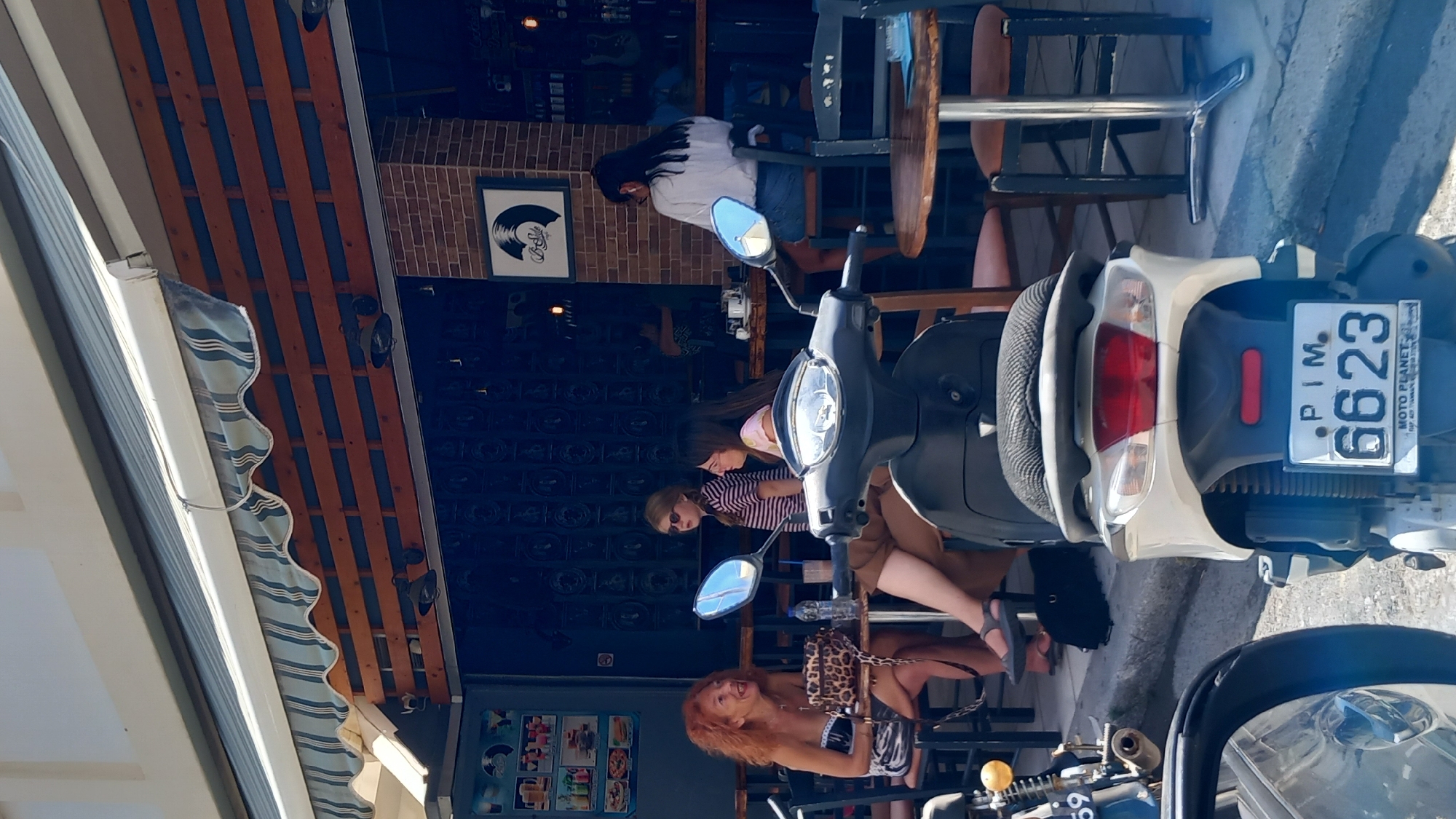 In the city of Kos, Greece, this image captures a vibrant outdoor cafe scene. Four individuals are seated at tables under a striped awning. On the left, a woman with vibrant red curly hair, wearing a strapless patterned top, engages in conversation while a leopard-print purse rests on the table. Adjacent to her, another woman, partially obscured, wears glasses and a light-colored top. A young girl with blonde hair, wearing a striped t-shirt and sunglasses, stands between them. Further to the right, a woman with long dark hair, wearing a white shirt and denim skirt, faces away from the camera, seated at a bar. In the foreground, a white scooter with the license plate "PIM. 6623" is parked, its reflection visible in a car's side mirror. The cafe's exterior features a wooden slat wall and exposed brick, with dark interior decor and posters visible through the open storefront. The overall atmosphere suggests a casual daytime gathering at a cafe.