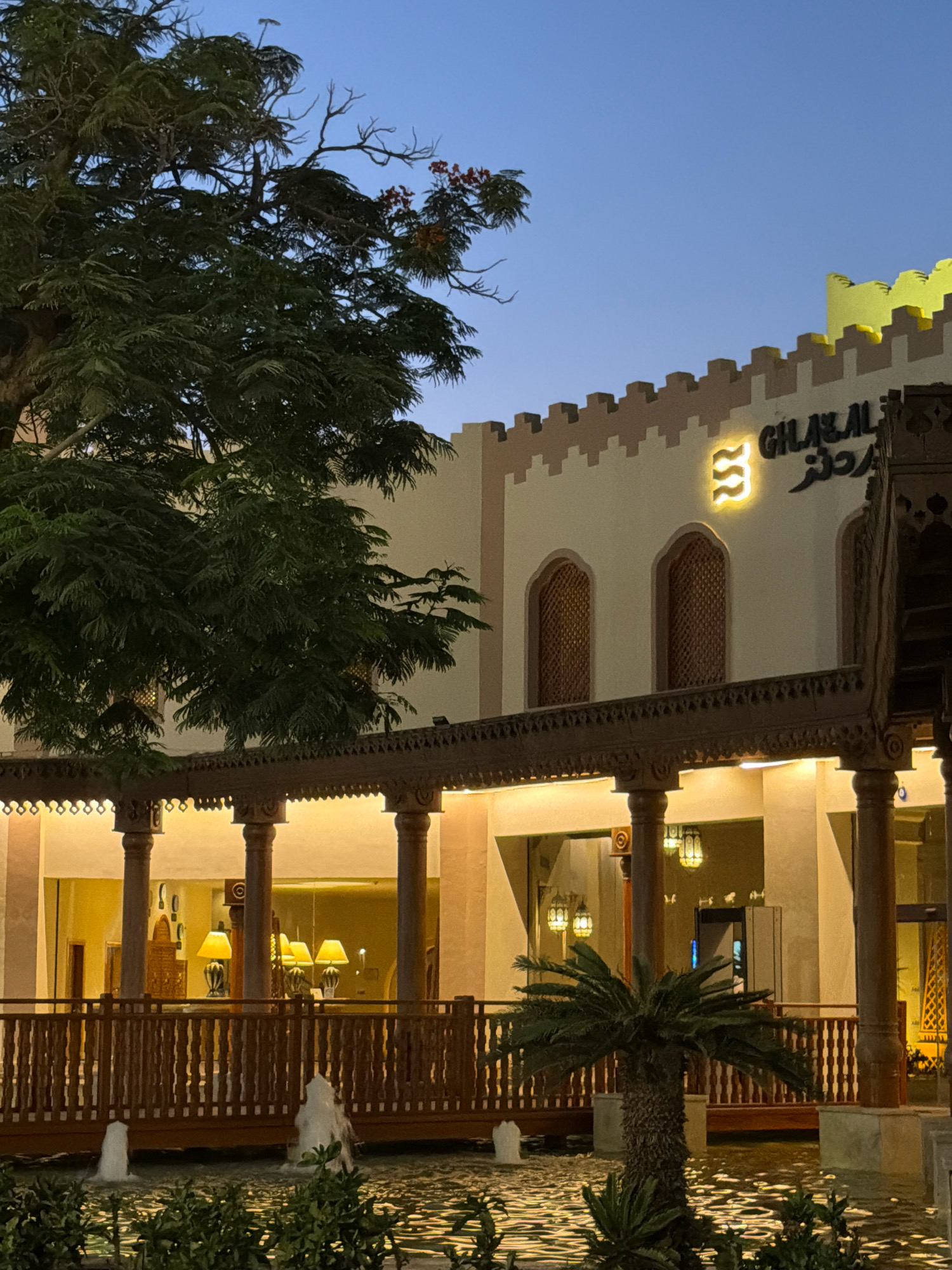 The image depicts the exterior of the Ghallazal restaurant in Sharm El Sheikh, Egypt, during twilight. A large tree with lush green foliage dominates the left side of the frame, its branches reaching towards the dusky blue sky. The restaurant building features a distinctive crenellated facade with a light beige color and brown accents. Above the entrance, the restaurant's name, "GHALLAZAL," is illuminated in Arabic and English, accompanied by a glowing logo.
A wooden walkway with ornate carvings and supporting pillars runs along the front of the building, offering a sheltered area with warm lighting from hanging lamps. In front of the walkway, a shallow water feature with active fountains adds a dynamic element to the scene. A small palm tree stands near the water, its fronds casting shadows. The overall atmosphere is tranquil and inviting, suggesting a sophisticated dining establishment in a resort setting. The lighting indicates that it is evening, with artificial lights illuminating the restaurant and its surroundings.