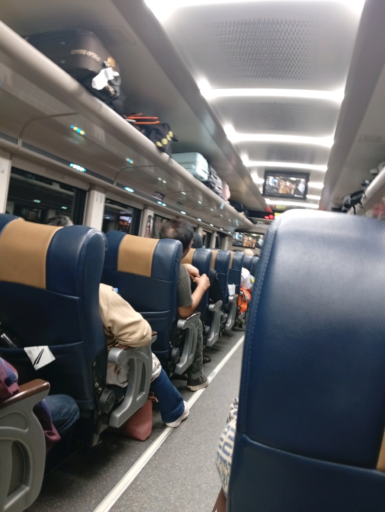 The image shows the interior of a passenger train, likely in Dukuhwidara, Indonesia, due to the context provided. The scene is viewed from the perspective of a passenger in a forward-facing seat, looking down the aisle. The seats are upholstered in dark blue with tan headrests. Several passengers are visible, some seated and others in the aisle. Luggage is stowed on overhead racks. Two small screens are mounted on the ceiling, displaying what appear to be people, suggesting entertainment or information displays. The lighting is artificial, with overhead fluorescent lights. The overall atmosphere appears to be that of a typical train journey, with passengers occupied with their own activities.