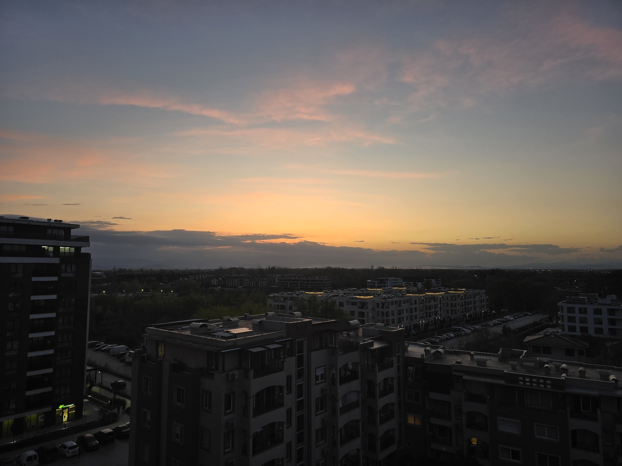 The image shows a cityscape at sunset. The sky is a gradient of colors, with orange and yellow near the horizon transitioning to pink and blue higher up. There are clouds visible in the sky.
Below the sky, there are buildings of varying heights and architectural styles. A tall, dark-colored building is on the left side of the image. There are other buildings in the midground, and they appear to be residential. There are also trees and vegetation visible between the buildings. Cars are parked on the streets. The location is in the city of Unknown, Unknown.