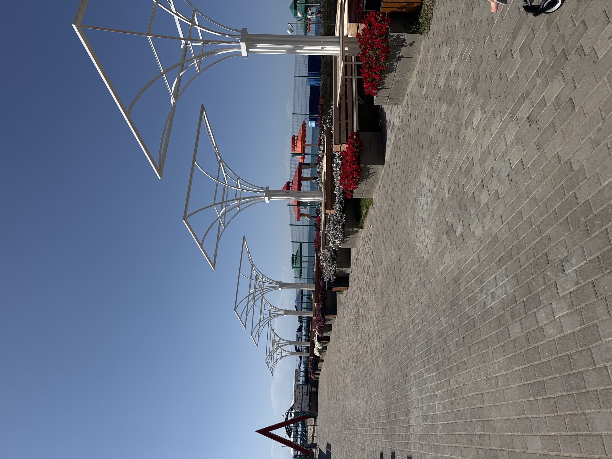 The image shows a paved walkway lined with planters containing red and white flowers. Several white, modern-style pergolas are spaced along the walkway, providing shade over wooden benches and tables. In the background, there is a view of a body of water, likely Lake Issyk-Kul, with a fence, more colorful umbrellas, and some structures that appear to be part of a resort or beach area. To the left, a red triangular arch structure is visible, and a person in a light-colored dress is walking away from the camera on the walkway. The sky is clear and blue. This scene depicts an outdoor recreational area, likely a promenade or seating area near a beach in Sary-Oy, Kyrgyzstan.