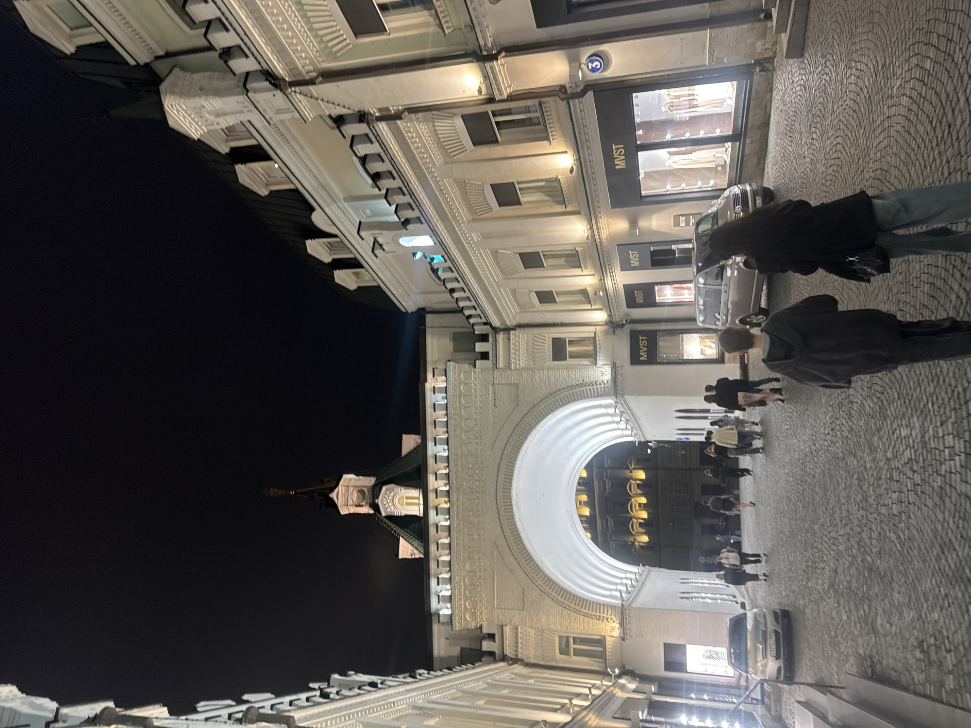 The image shows an evening scene in Moscow, Russia. The location is likely near Red Square, identifiable by the archway and a glimpse of the Spasskaya Tower with its illuminated green roof and red star. The sky is dark, indicating nighttime. Several people are walking on a cobblestone street. Street lights and building lights illuminate the area. A car is parked on the right side of the frame. The building on the right side of the street has storefronts labeled "MVST". A building across the street has a storefront with number three on it.