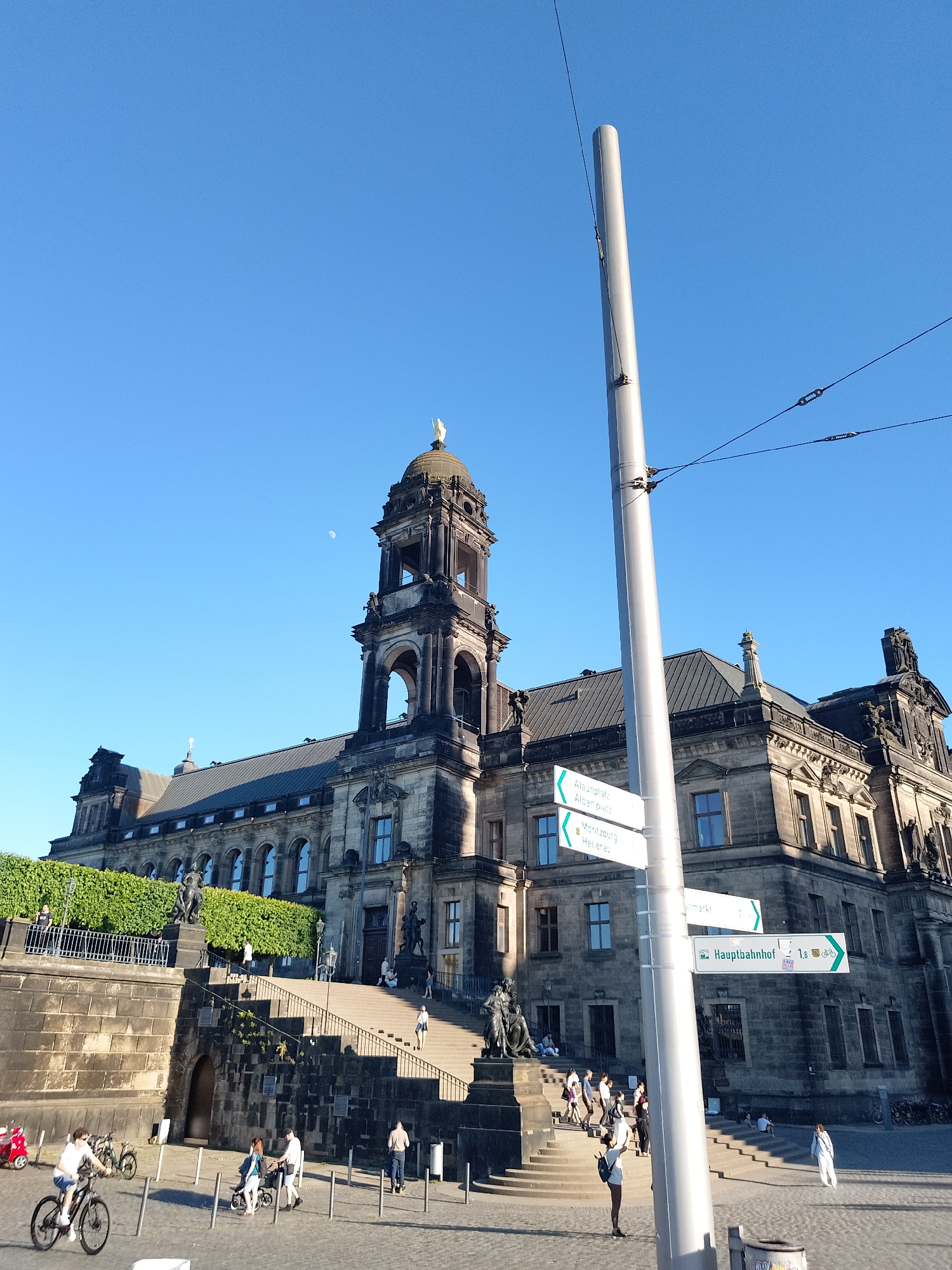 The image shows a large, ornate stone building, likely a historical or governmental building, under a clear blue sky. A tall tower topped with a golden statue is visible. The building has multiple windows and architectural details.
A street lamp post with several direction signs obscures part of the building. One sign points toward "Hauptbahnhof" (Main Train Station).
A wide stone staircase leads up to the building, with people walking up and down. Several people are standing, sitting, or walking in the plaza at the base of the staircase. Some are on bicycles.
The location is likely Dresden, Germany, based on the architectural style and the Hauptbahnhof sign.