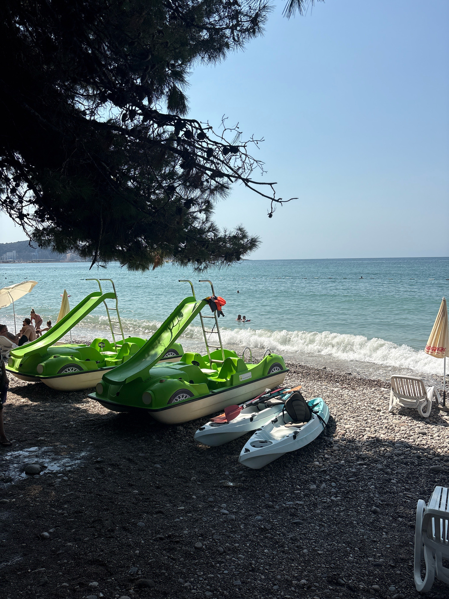 The image depicts a pebbled beach on a sunny day, likely in Bar, Montenegro. In the foreground, two green pedal boats with slides are parked on the shore, side-by-side. Two kayaks are placed in front of the pedal boats. The sea, with moderate waves, stretches to the horizon under a clear blue sky. A large pine tree casts a shadow over the left side of the scene, with its branches visible at the top. In the background, people are visible in the water and on the beach, along with white beach umbrellas and lounge chairs.