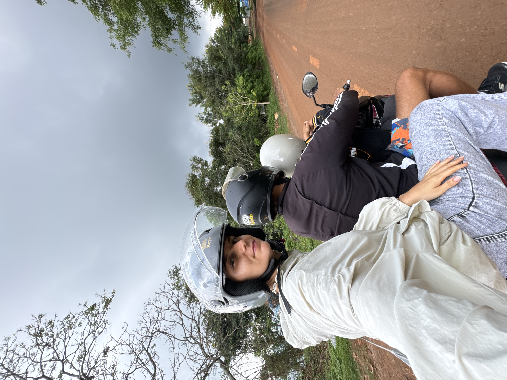 Three people are riding a motorcycle on a dirt road. The person in front, a woman, is wearing a white hooded jacket and grey patterned pants. She is looking back at the camera and smiling. She has a clear helmet on. The person in the middle is wearing a black jacket and a black helmet. The person in the back is wearing a light grey helmet. The motorcycle is moving forward on a red dirt road that is lined with trees. The sky is cloudy. The location is Nao Sẻ, Vietnam.