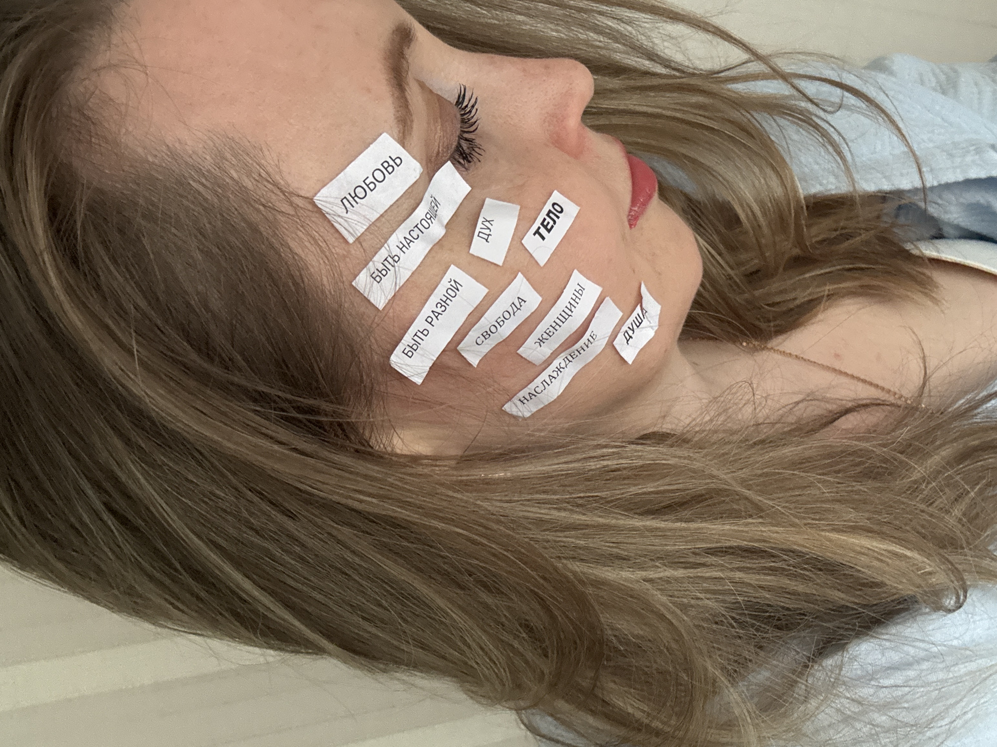 A close-up, angled shot shows a woman lying down, possibly indoors, in Chaikovsky, Russia. Her face is presented in profile, with her eyes closed or heavily lidded, and she has pronounced dark eyelashes. Her lips are slightly parted, highlighted with red lipstick. Her long, light brown hair, interspersed with blonde highlights, frames her face and flows across a light-colored, soft surface, likely a bed or pillow. A delicate gold chain is visible around her neck.
The notable activity involves small, rectangular pieces of white paper or tape affixed to her face, each bearing a Russian word. This arrangement suggests a symbolic or introspective exercise.
The visible text on the papers, from top to bottom and left to right, includes:
* "ЛЮБОВЬ" (LYUBOV) - Love
* "БЫТЬ НАСТОЯЩЕЙ" (BYT' NASTOYASHCHEY) - To be real / To be authentic
* "ДУХ" (DUKH) - Spirit
* "ТЕЛО" (TELO) - Body
* "БЫТЬ РАЗНОЙ" (BYT' RAZNOY) - To be different / To be diverse
* "СВОБОДА" (SVOBODA) - Freedom
* "ЖЕНЩИНЫ" (ZHENSHCHINY) - Women
* "НАСЛАЖДЕНИЕ" (NASLAZHDENIYE) - Pleasure / Enjoyment
* "ДУША" (DUSHA) - Soul
The overall impression is one of calm and self-reflection, with the words pointing to significant aspects of personal experience and identity. The lighting is soft and even, suggesting an indoor environment without specific time-of-day cues.