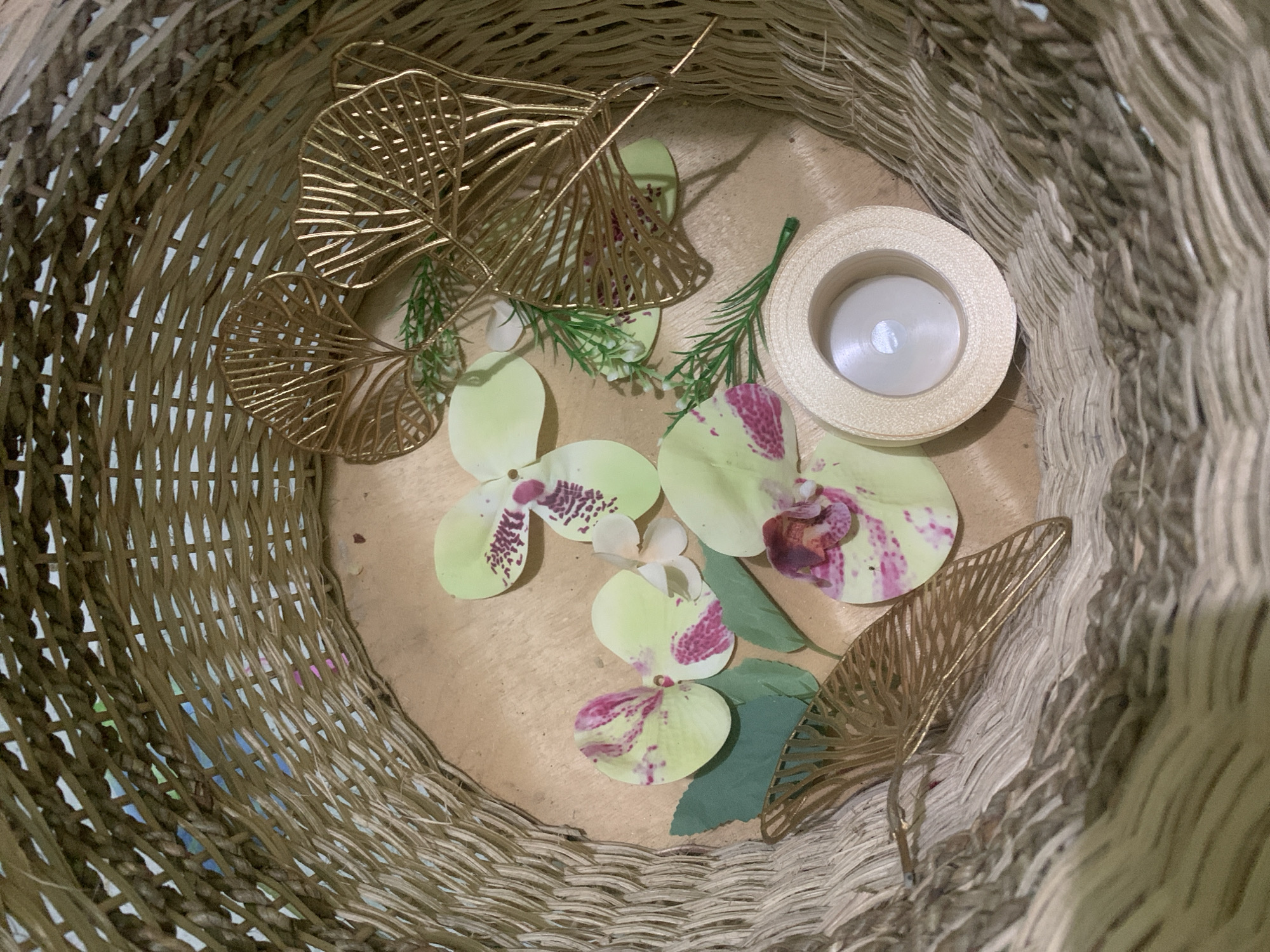 Inside a woven basket, a collection of decorative items is arranged on a light brown wooden surface. The items include several light green orchid petals with pink speckles, two small green artificial leaves, and three ornate gold leaf-shaped embellishments. Additionally, a small roll of cream-colored ribbon and a sprig of artificial greenery are present. The basket itself is made of a coarse, light brown woven material. The overall scene is a still life arrangement, with no people or animals visible. The context of Kuala Pembuang, Indonesia, is not discernible from the visual information provided in the image.