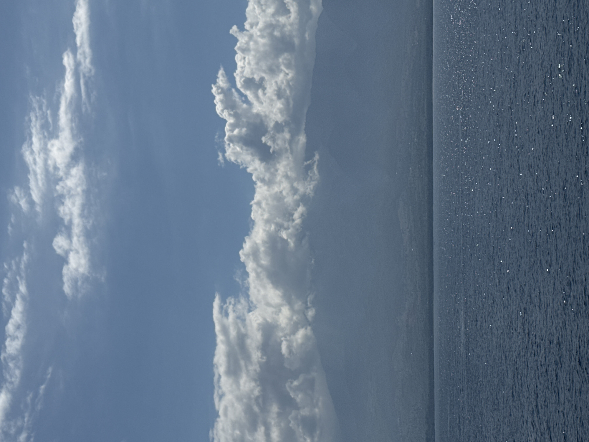 An overhead shot of a body of water with a line of mountains in the distance. The mountains are covered in a thin layer of haze. The sky is a clear blue with a scattering of white clouds. The water is a dark blue with small white highlights from the sun. The image is taken from a slightly elevated angle, suggesting the vantage point of a boat or a high building.