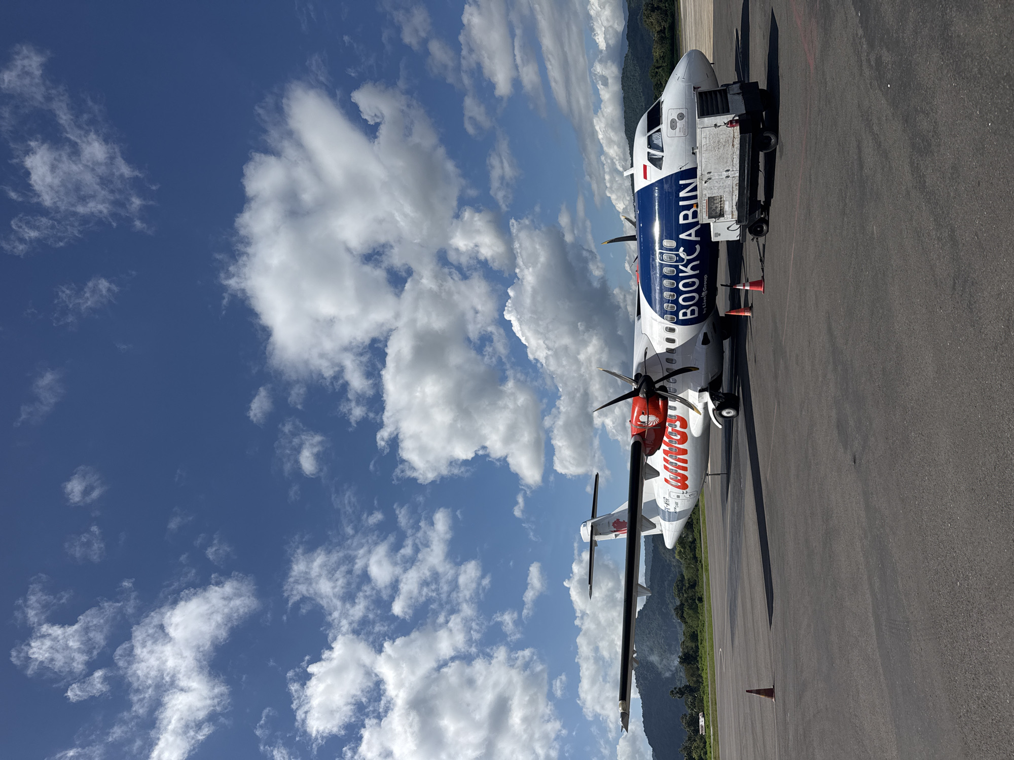 The image shows two planes parked on an airport tarmac under a partly cloudy sky. One plane has "BOOKCABIN" written on the side, and another one has "Wings Air" written on its tail. The location is likely in or near Labuan Bajo, Indonesia, based on the airline and general environment.