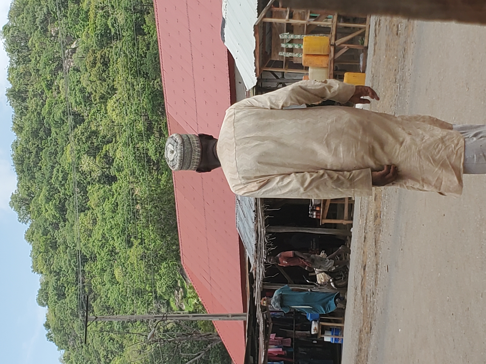 The image shows a street scene in Mararaba, Nigeria. A man in traditional attire, wearing a patterned cap and a light-colored robe, walks away from the camera. Behind him, a street with small shops or stalls lines the road. The structures appear to be made of wood and corrugated metal, with red or grey roofs. Various goods are displayed for sale. On the opposite side of the street, a steep, green hillside covered in lush vegetation rises sharply. Power lines run along the street.