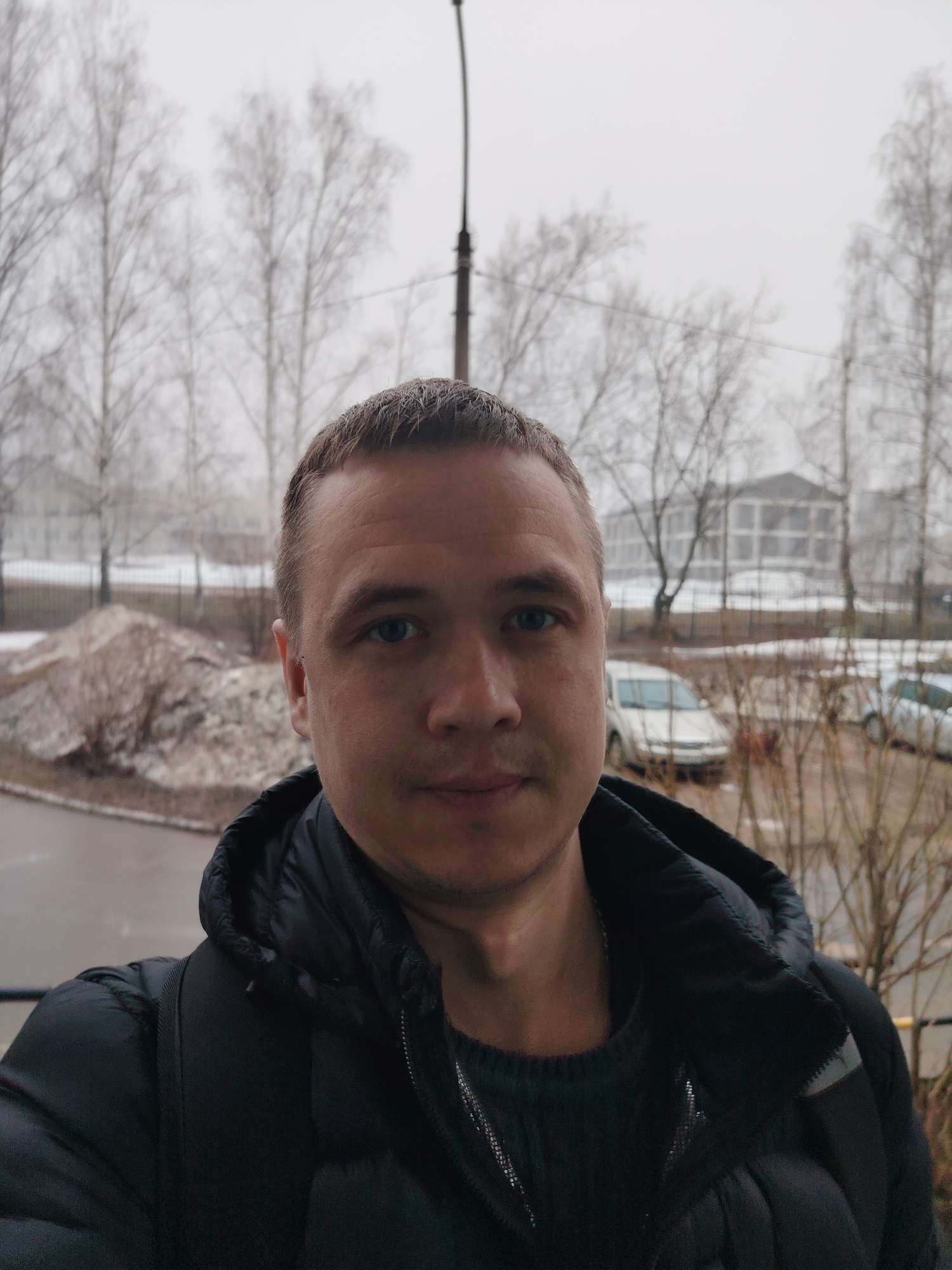 A man is taking a selfie in front of a snowy cityscape. He has short brown hair and blue eyes, and he is wearing a black puffer jacket over a dark sweater. Behind him is a street with parked cars, bare trees, and a building in the distance. The image appears to be taken on a balcony or raised platform. The weather is overcast and snowy. The location is Chaikovsky, Russia.