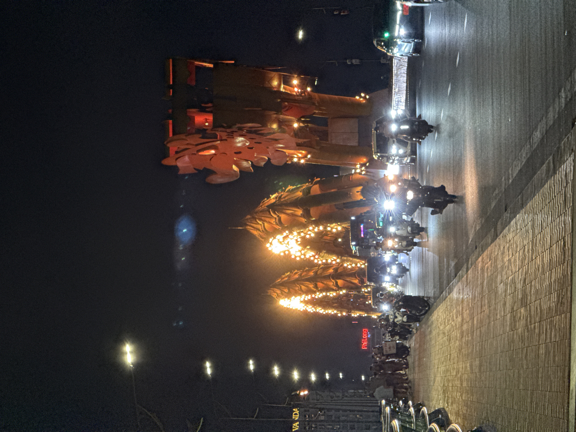 This image captures a nighttime scene on the Dragon Bridge in Da Nang, Vietnam. The iconic bridge, a symbol of the city, is illuminated with a vibrant orange glow, highlighting its dragon-like structure. The bridge spans a waterway, and traffic, primarily consisting of motorcycles and some cars, moves across it. The road is wet, reflecting the lights of the vehicles and streetlights. On the left side of the image, a pedestrian walkway paved with brick is visible, with several people strolling along it. Some of these individuals are gathered around a sign. The overall atmosphere is lively, with the city lights creating a dynamic and colorful urban landscape.