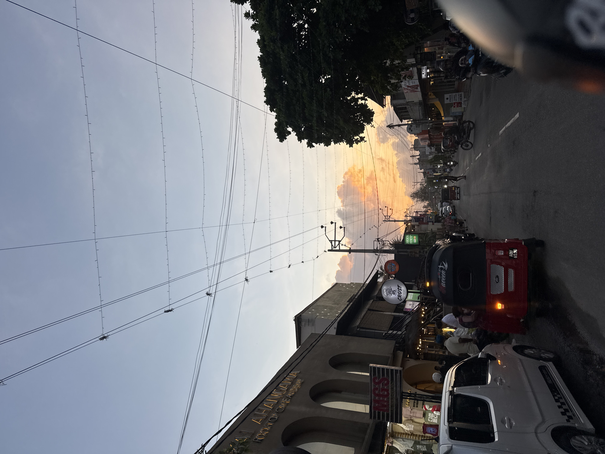 The image depicts a street scene in Ahangama, Sri Lanka, during sunset. Ahangama is a coastal town known for its surfing spots and laid-back atmosphere. The sky is filled with hues of orange and pink as the sun sets, casting a warm glow on the surroundings.
The street is lined with buildings that appear to be shops and restaurants, with signs such as "AL AHANGAMA ROOFTOP CAFE AND BAR," "MGS FASHION," "CRUST," and "HOTEL DE," visible. The architecture suggests a mix of modern and traditional styles.
Various modes of transportation are present, including a red tuk-tuk, a white car, and several motorcycles. People are seen walking on the sidewalks and riding the vehicles, suggesting a busy evening in the town.
Overhead, a network of power lines crisscrosses the sky, with some appearing to be adorned with string lights. A large, leafy tree on the right side of the image adds a natural element to the urban landscape. The overall impression is that of a vibrant and active street in a coastal town at dusk.
