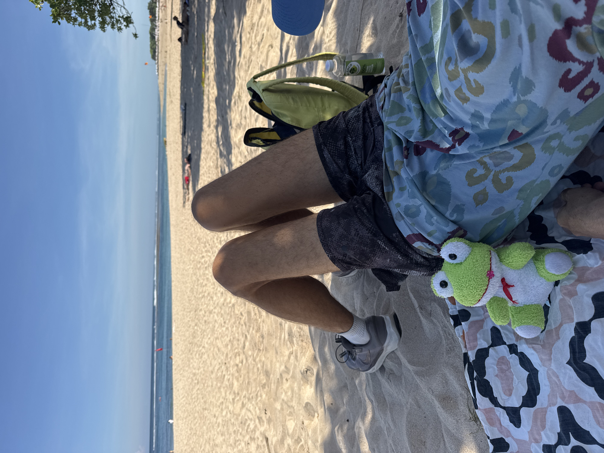 A person is sitting on a sandy beach in Kuta Selatan, Indonesia, overlooking the ocean. The person is wearing dark shorts, a patterned shirt, and gray sneakers with white socks. To the person's right, a stuffed frog toy rests on a patterned blanket. A blue hat and a green bag are also visible nearby. The background shows a clear blue sky, the ocean, and the beach stretching into the distance with trees and other people visible.