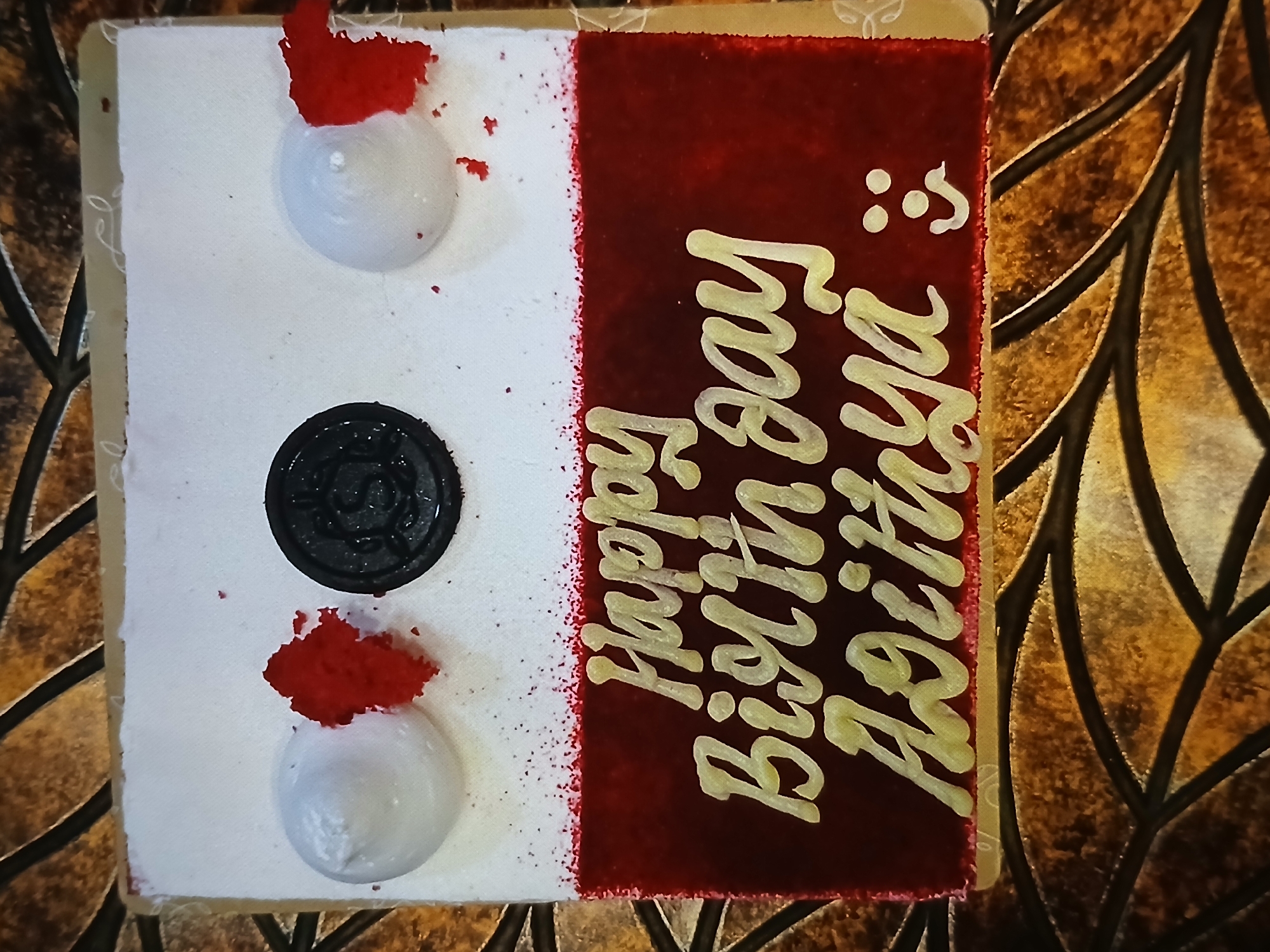 A square cake is positioned on a surface with a patterned background. The left side of the cake is white with three white frosting mounds, with red crumbles near two of them. A black disc, possibly an Oreo cookie, is centrally located on the white frosting. The right side of the cake is red and features "Happy Birthday Achiya" written in a golden color. The background appears to be a reflective surface with leaf-like patterns. There is no indication in this image of this being from Bengaluru, India.