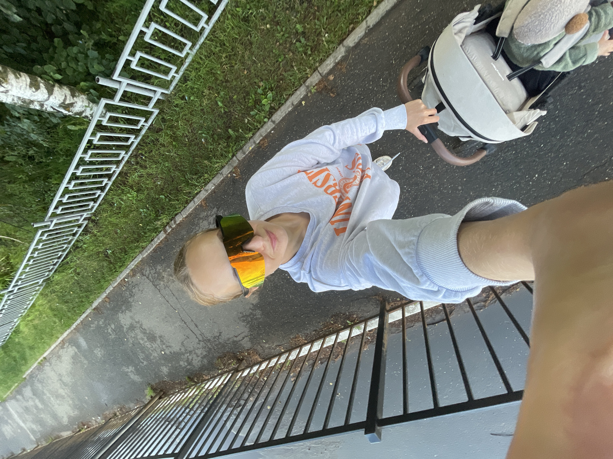 The image shows a woman in athletic sunglasses and a light-colored sweatshirt with orange lettering, holding the handle of a stroller. She is standing on a paved walkway next to a grassy area bordered by a white fence. The scene appears to be outdoors, possibly in a park or residential area. There is a dark metal fence to the left of the walkway. The woman is holding her phone, suggesting she is taking a selfie or recording a video. The stroller is light-colored with brown handles. The overall impression is of a casual outdoor activity, possibly a walk with a child in the stroller. The location of Подшивалово, Russia is not directly indicated by any visual cues in the image.