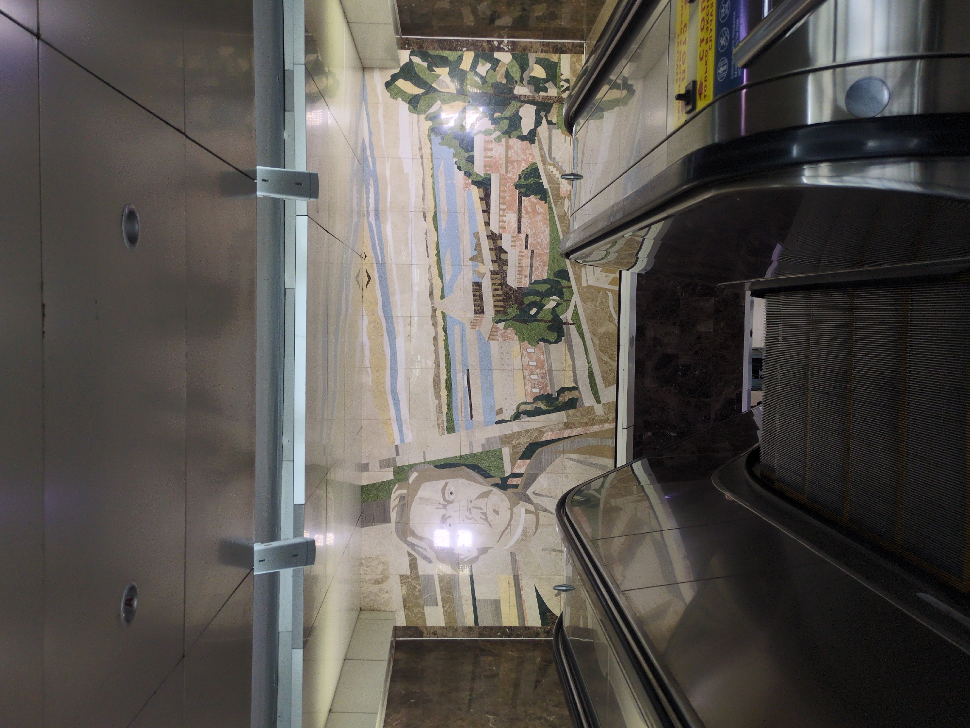 The image shows a wide view of an escalator in a subway station. The escalator is dark gray and metallic, with ribbed steps. The walls of the station are covered in a large mosaic mural. The mosaic depicts a portrait of a man on the left side, and a landscape scene on the right. The landscape includes a river, buildings, trees, and a sky with birds. The overall environment appears to be indoors, with a tiled ceiling and lighting fixtures visible. There are no people visible in the image. Based on the architecture and the mosaic style, this is likely a station in the Nizhny Novgorod Metro, Russia.