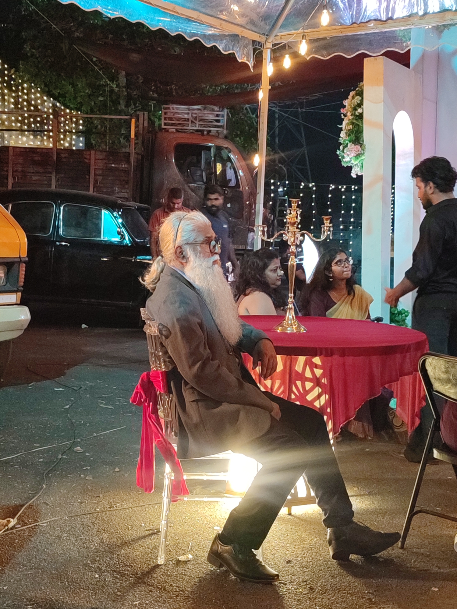 The image captures an evening gathering in Kalamassery, India, held outdoors under a translucent canopy or tent illuminated by string lights. The setting appears to be a casual yet decorated event, possibly a reception or party, taking place at night.
In the foreground, an older man with a distinctive long white beard, a ponytail, and spectacles is seated on a clear plastic chair adorned with a red fabric ribbon. He wears a dark suit jacket over a white shirt and dark trousers, with a light source illuminating his lower body. He is positioned to the left, observing the scene before him.
Behind the man, a round table draped in a vibrant red tablecloth features a golden candelabra centerpiece. Two women are seated at this table. One woman, wearing a light-colored saree, looks up towards a standing man on the right side of the frame, who is dressed in a dark shirt and trousers, seemingly engaged in conversation. Another woman is seated next to her. The tablecloth is lit from underneath, creating a warm glow and highlighting decorative patterns on the fabric.
The background reveals several vehicles: a classic black sedan (likely a Hindustan Ambassador), a portion of a yellow van, and a larger brown truck parked behind them. String lights are visible along a structure or fence in the distant background, adding to the festive ambiance. The ground is dark, possibly asphalt or packed earth, with some debris visible. There is no visible text in the image.