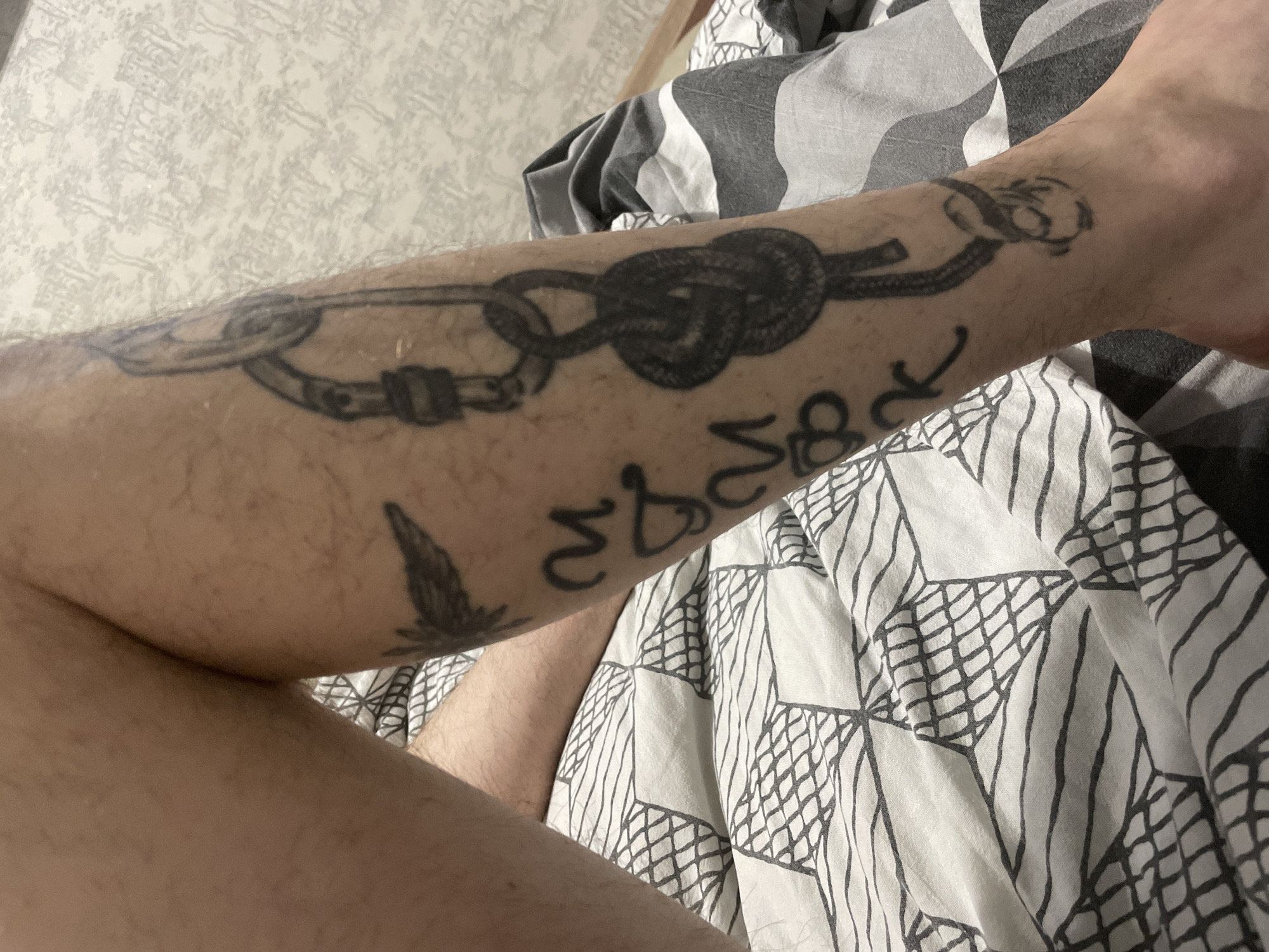 The image shows a fair-skinned forearm with a tattoo. The tattoo appears to depict a climbing rope secured to a carabiner, another climbing knot, potentially a figure eight, and a symbol that might be related to climbing. There is also a tattoo of a feather near the elbow crease. The arm is resting on a bed with patterned sheets in shades of gray and white. The background is blurred, suggesting a domestic setting. The location is provided as Sarapul, Russia; however, this cannot be verified from the image itself.