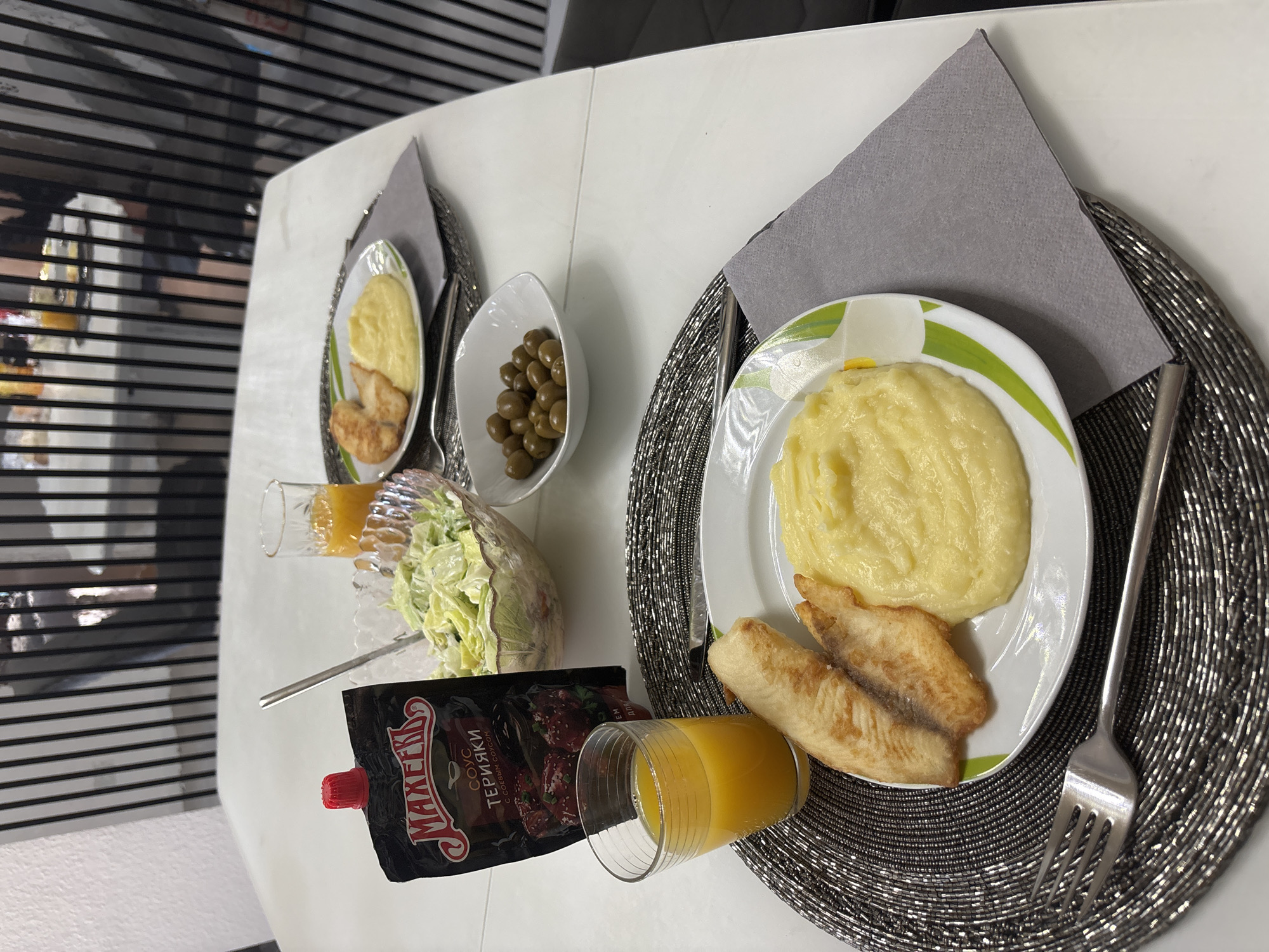 Two place settings are visible on a white table. Each setting includes a plate with mashed potatoes and fish fillets, a glass of orange juice, and a gray napkin. One setting also has a fork, while the other has a fork and a knife. A bowl of salad and a small bowl of olives are on the table, along with a pouch of teriyaki sauce. The background features a black and white striped vertical partition. The scene appears to be indoors, likely in a dining area. No people are visible.