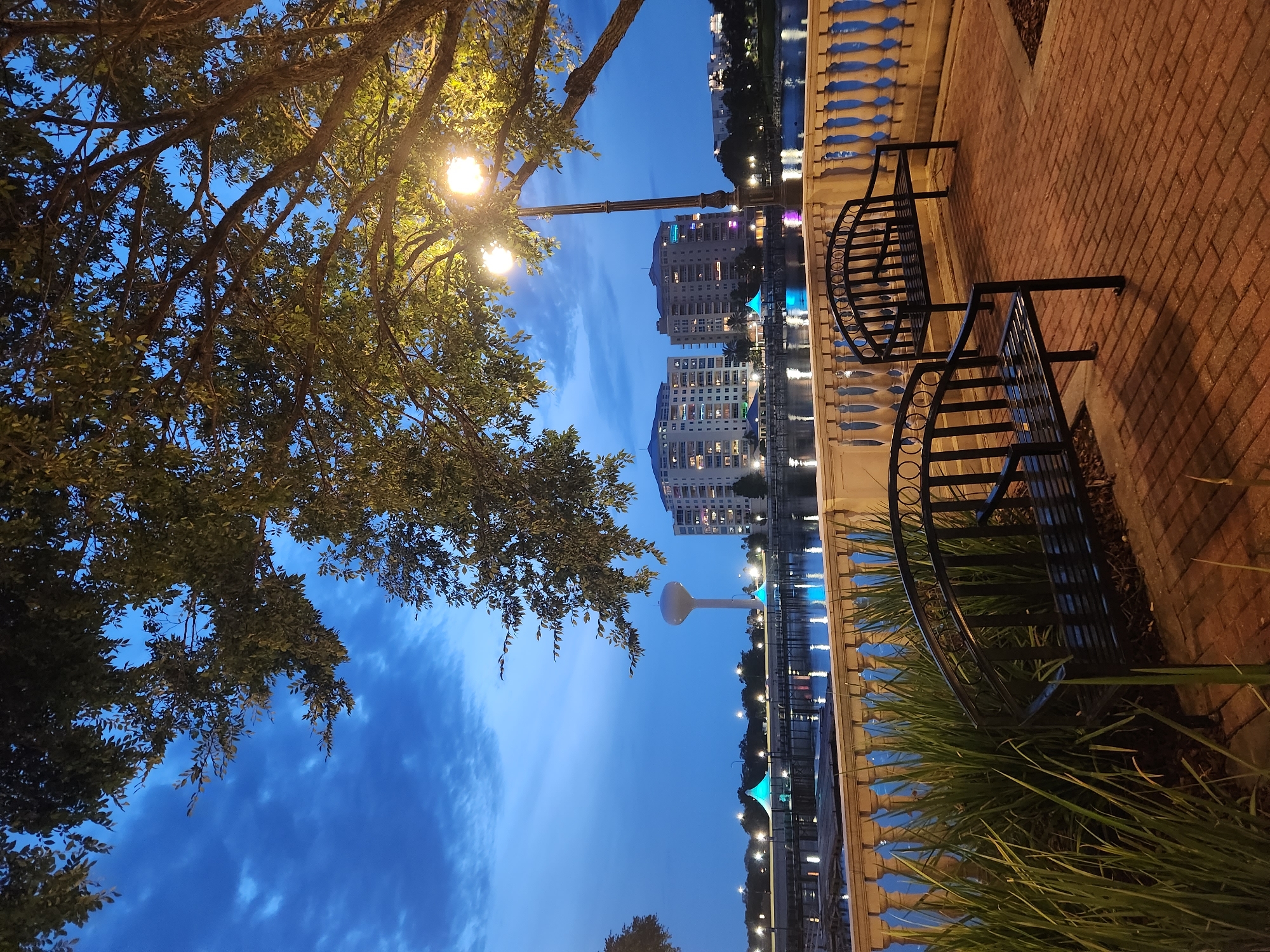 The image depicts a waterside promenade at dusk in Altamonte Springs, Florida. Two black metal benches with ornate backs are situated on a brick-paved area, facing a body of water. A decorative balustrade separates the promenade from the water. Across the water, a row of lights illuminates a walkway, with two teal-colored tents visible. Further in the background, a large white water tower stands, flanked by modern apartment buildings with illuminated windows. A streetlamp with two bulbs casts a warm glow over the benches. Lush green foliage, including tall grass and the branches of a large tree with illuminated leaves, frames the scene. The sky is a deep blue, indicative of twilight, with scattered clouds. The overall impression is one of a tranquil public space by the water as evening descends.