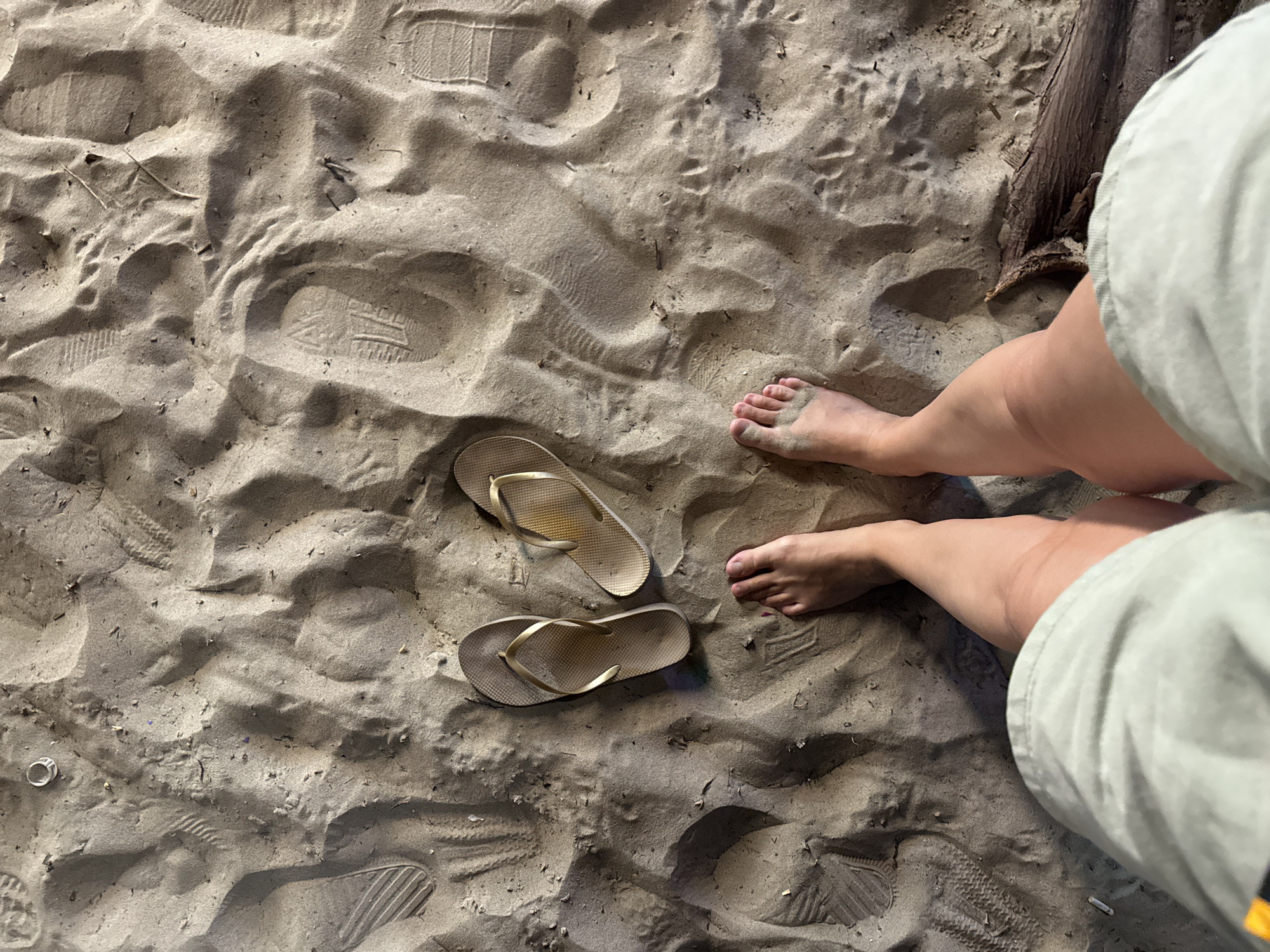 The image depicts a top-down view of a sandy beach, likely located in Da Nang, Vietnam, given the context. In the foreground, a person's bare feet are visible, with their toes pointing upwards. The feet are slightly tanned and appear to be on the sand. To the left and right of the feet are two flip-flops, one gold and one beige. Both flip-flops are positioned on the sand, with the gold one on the left and the beige one on the right, slightly further away. The sand itself is textured with numerous footprints, some appearing to be from shoes with a distinct tread pattern, while others are less defined. There are also a few small pieces of debris scattered on the sand, including a twig and what might be a cigarette butt. In the upper left corner, a ring is visible on the sand. The overall scene suggests a casual setting on a beach, possibly after a walk or during a break.