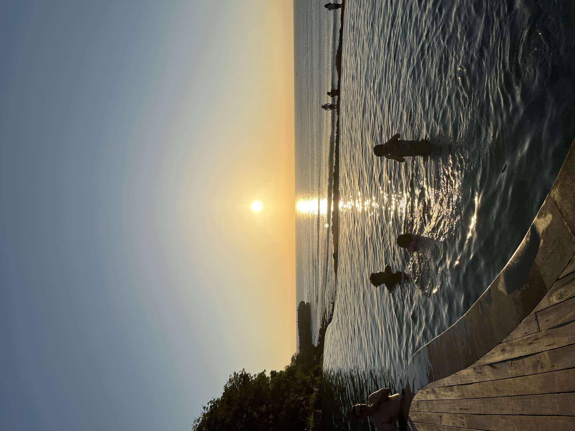 An infinity pool with a wooden deck is in the foreground, overlooking the ocean at sunset. Several people are in the pool, with some standing and others swimming. In the background, the sun is setting over the ocean, casting a golden glow on the water. A rocky outcrop with a few people standing on it is visible in the distance, along with a tree-covered cliff face on the left side of the image. The scene is located in the city of Kuta Selatan, Indonesia.