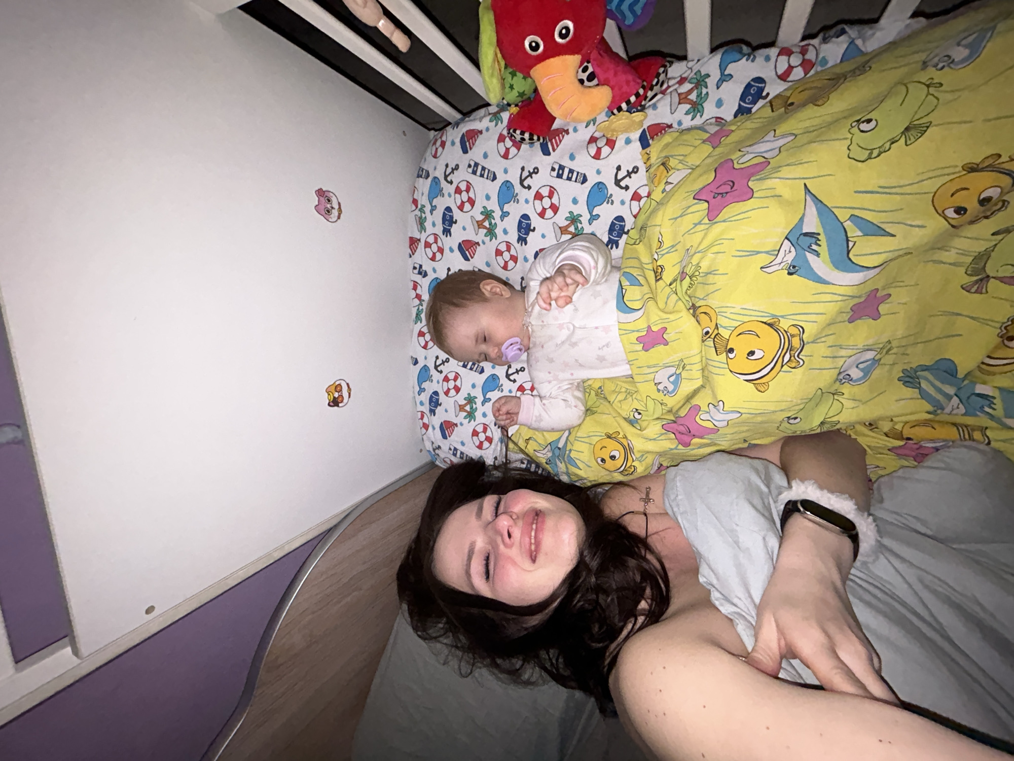 This indoor image captures a tender moment between a woman and a sleeping baby, likely in a bedroom or nursery in Rusalevka, Russia. The perspective is from above, suggesting a selfie or a shot taken from a raised position.
The woman, with dark, wavy hair, is lying on her back, smiling broadly and looking towards the camera. She wears a light-colored top and a necklace with a small cross pendant. A smart band with a fluffy white strap is visible on her left wrist. She appears relaxed and content.
Next to her, in a crib, a baby with light hair sleeps soundly on its side, a purple pacifier in its mouth. The baby is dressed in a white long-sleeved outfit. The crib's fitted sheet features a cheerful pattern of anchors, lifebuoys, and blue whales. A colorful plush toy, resembling a red elephant, hangs above the baby from the crib bars. The baby is partially covered by a bright yellow blanket adorned with various cartoon fish and starfish, reminiscent of characters from "Finding Nemo."
The background reveals a plain white wall with a few small, colorful cartoon animal stickers, including a pink pig-like face and an orange creature. A section of purple wall is visible below the white wall, along with some wooden furniture. The lighting appears dim, suggesting the photo was taken at night or in a softly lit room. There is no visible text in the image.