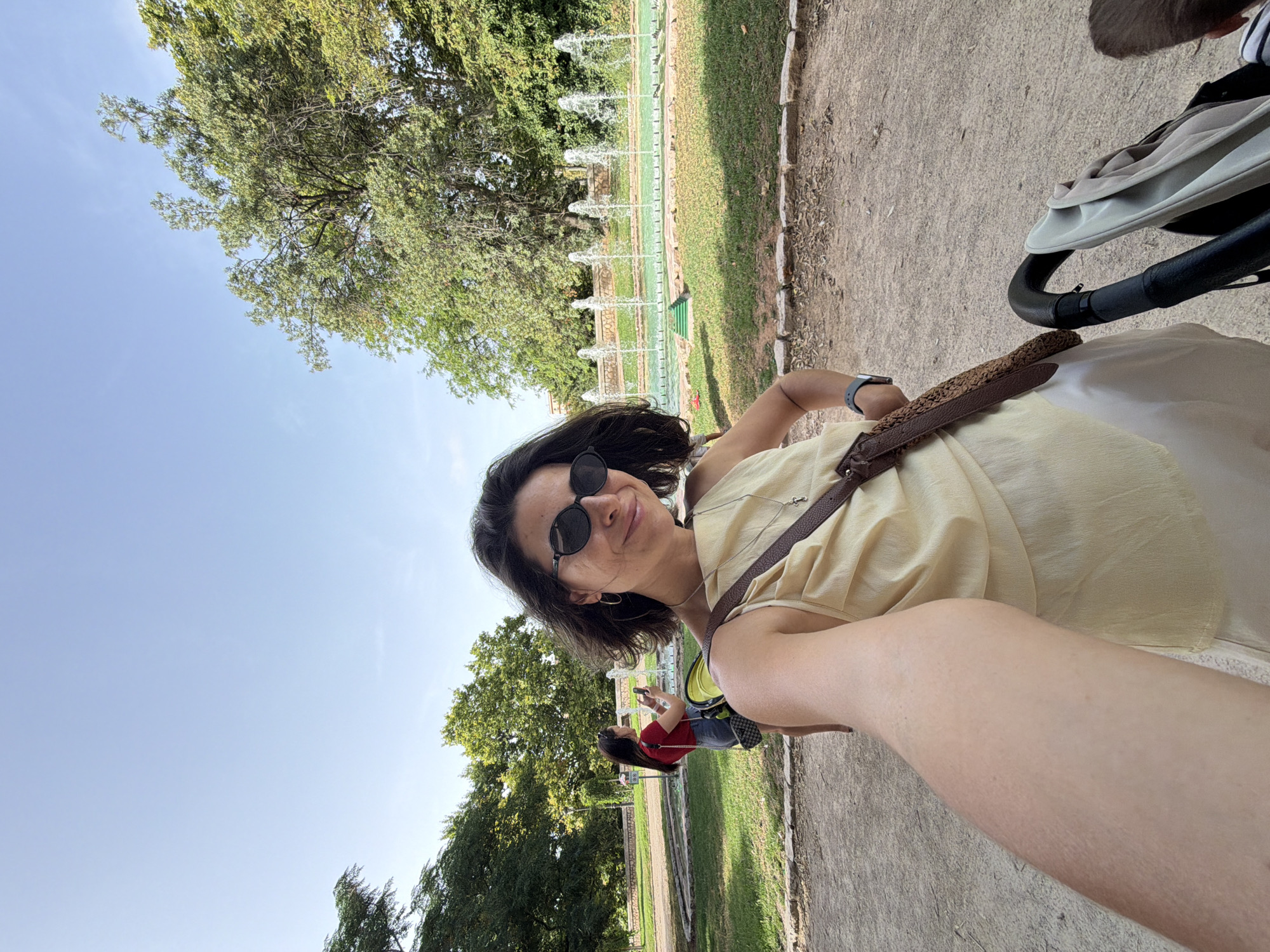 The media file is a selfie taken outdoors in what appears to be a park or garden. The main subject is a woman with dark, shoulder-length hair, wearing sunglasses and a light yellow top. She is smiling and looking at the camera. A dark-brown woven strap is visible across her shoulder, and she has a watch on her left wrist.
In the background, there is a long fountain with multiple jets of water spraying upwards. Beyond the fountain, there is a well-maintained lawn and trees with lush green foliage. The sky is clear and blue, suggesting it is daytime, likely sunny. The ground in the foreground is a gravel or dirt path. A stroller is partially visible to the right of the woman, suggesting she might be with a child or pushing one. Another person wearing a red top is visible in the mid-ground, looking at their phone. The overall scene evokes a relaxed and pleasant outdoor atmosphere.