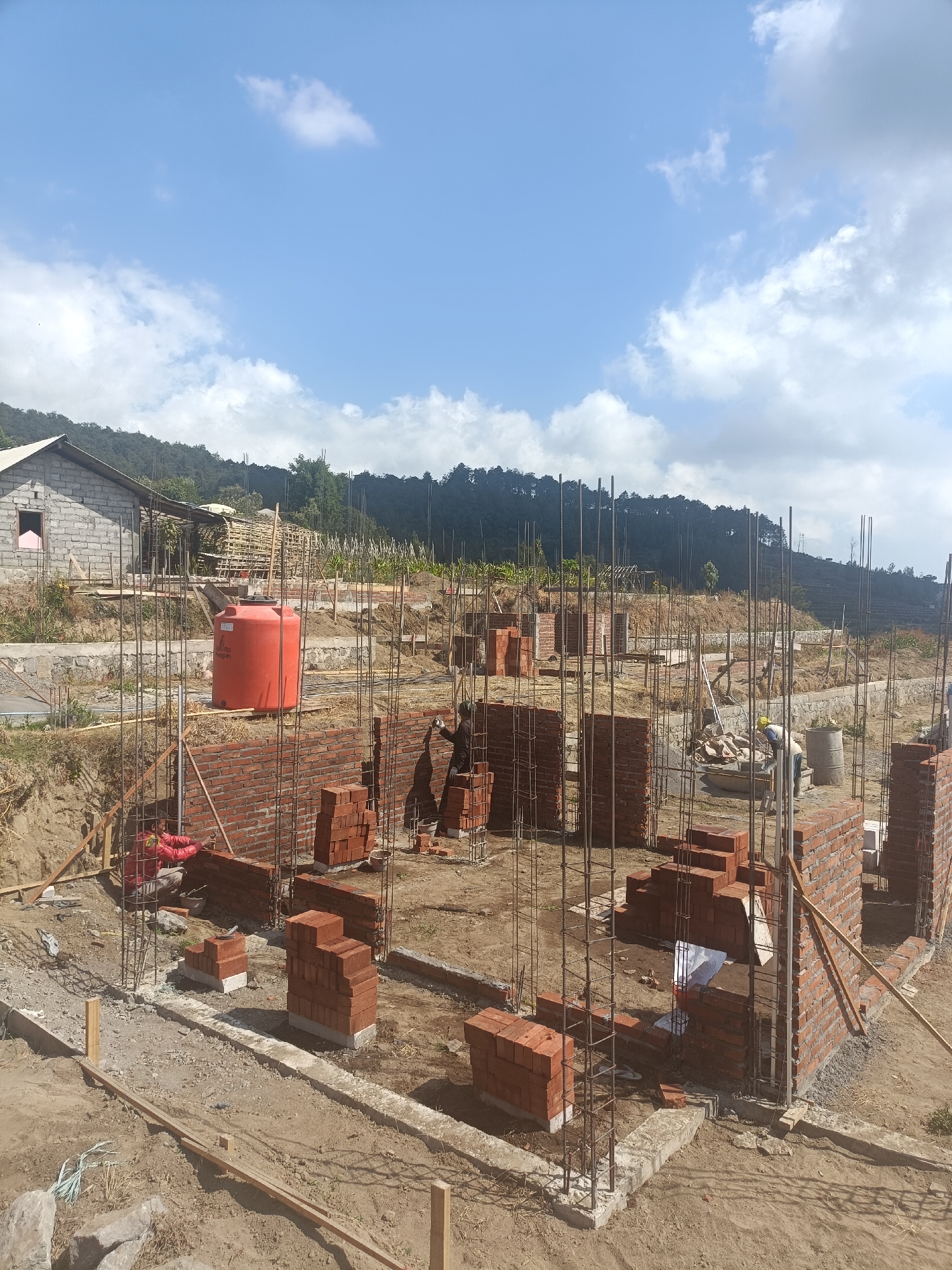 The image depicts a construction site in Kopeng, Indonesia, under a bright blue sky with scattered clouds. The foreground shows foundations and brick walls in various stages of construction. Numerous steel reinforcing bars are visible, protruding from concrete bases, indicating structural work in progress. Stacks of red bricks are neatly arranged, ready for use.

In the mid-ground, a large orange water tank stands prominently. A person in a red jacket is actively laying bricks on a wall, wearing white gloves. Another individual, wearing a yellow hard hat, is seen working near a concrete mixer and a barrel. A partially constructed building with a concrete block exterior is situated in the background, to the left, with a glimpse of a pink-framed window.

The surrounding environment is hilly and covered with vegetation, with trees and a treeline visible on the slopes in the distance. The ground is a mix of dirt and gravel, typical of an active construction area. The overall scene conveys a sense of ongoing development and labor.