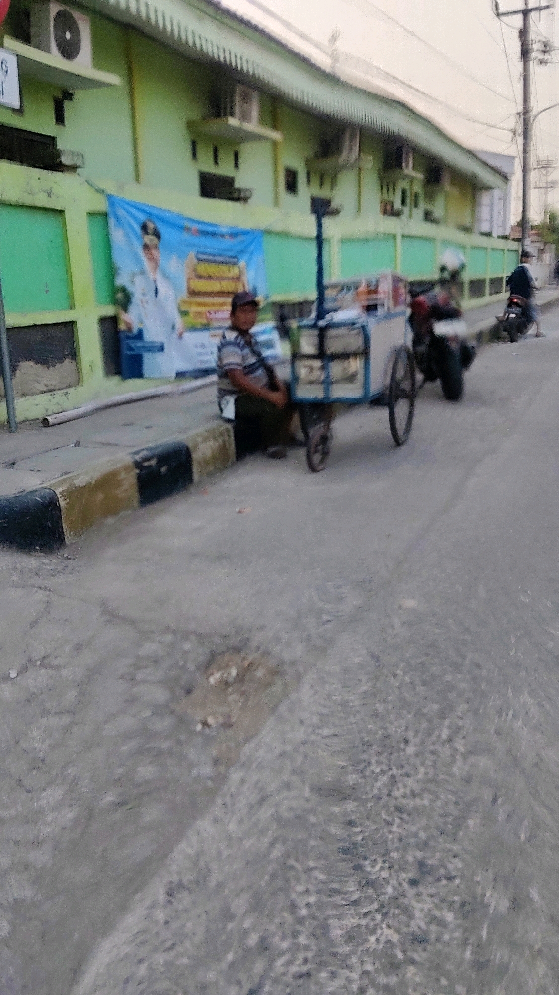 A person wearing a striped shirt and a dark cap is seated next to a food cart on the side of a street in Mekarmukti, Indonesia. The food cart, with a light-colored body and blue trim, has two wheels visible. In the background, a light green building with several air conditioning units is present, along with a blue banner displaying an image of a person in a white uniform. Another person is visible on a motorcycle further down the street. The pavement in the foreground shows signs of wear and tear, with some debris scattered on it. The scene appears to be outdoors, possibly in a commercial or residential area.