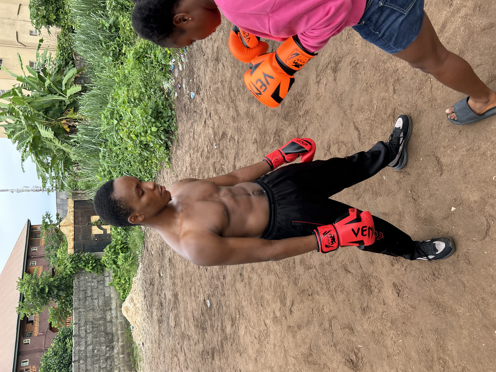 In Ihiagwa, Nigeria, two individuals, a shirtless young man with defined abdominal muscles and a young woman wearing a pink sweatshirt and denim shorts, stand on a dirt ground. Both are equipped with boxing gloves; the man wears red gloves and the woman wears orange gloves. The man is positioned in the foreground, facing left with his head slightly bowed, while the woman is in the background, facing right. Behind them, a weathered brick wall separates the dusty area from a lush green environment with dense vegetation, including banana trees. Further back, multi-story buildings with visible windows and balconies are present. A rusty, ornate gate is set into the wall. The overall scene suggests an outdoor boxing practice or training session in a semi-urban or rural setting.