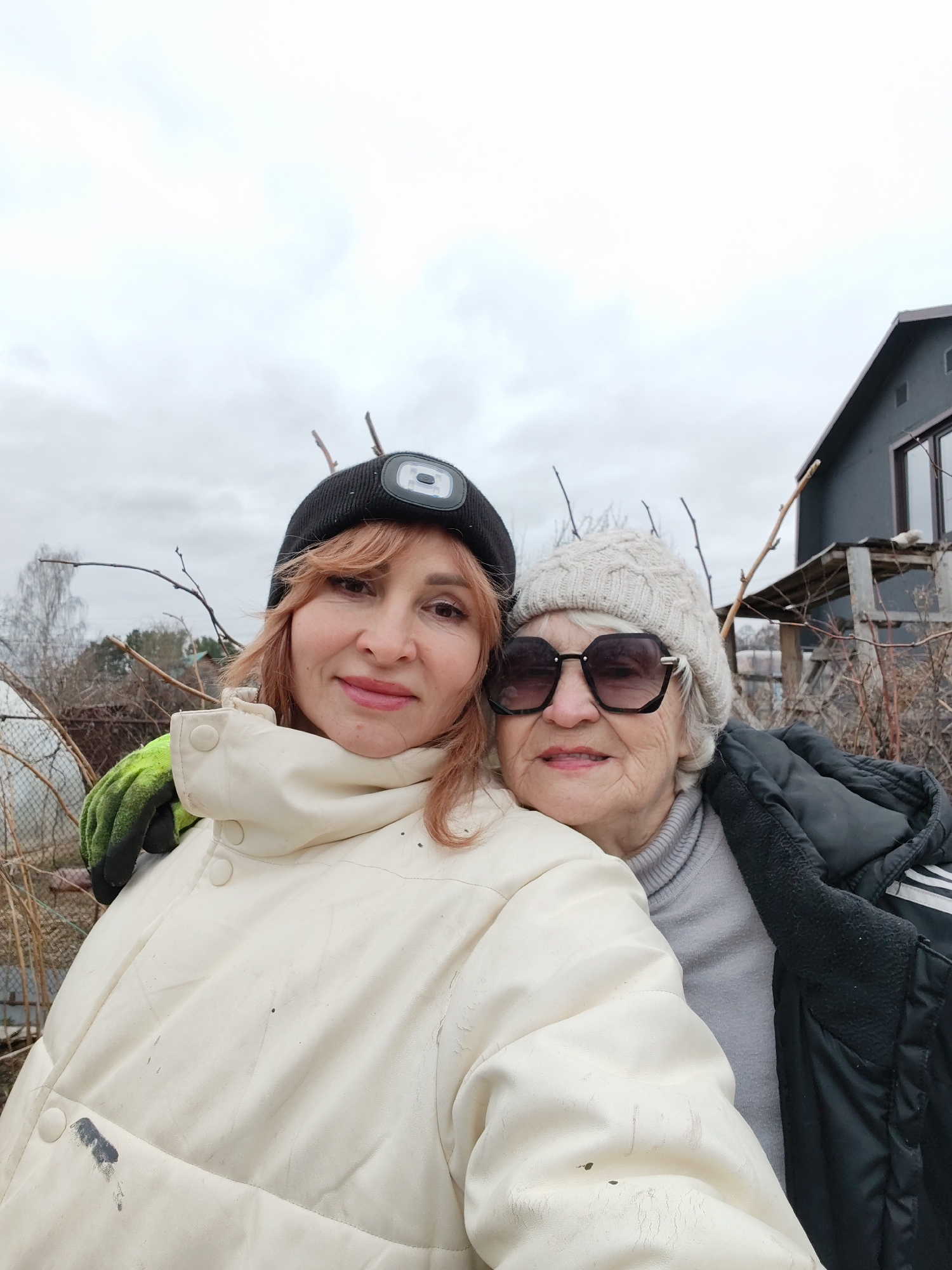 Two women are closely positioned in a selfie-style photograph, appearing outdoors in an environment that suggests they are in a garden or yard. The younger woman, on the left, has red hair and is wearing a black beanie with a headlamp attached, a cream-colored jacket, and a green gardening glove. The older woman, on the right, has short white hair, wears a light-colored knitted hat, large sunglasses, and a gray turtleneck sweater under a black jacket. Behind them, there is foliage with bare branches, a wire fence, and a dark gray or black building in the background. The sky is overcast, suggesting a cool, possibly winter or early spring day. The photograph appears to be taken in Chaikovsky, Russia.