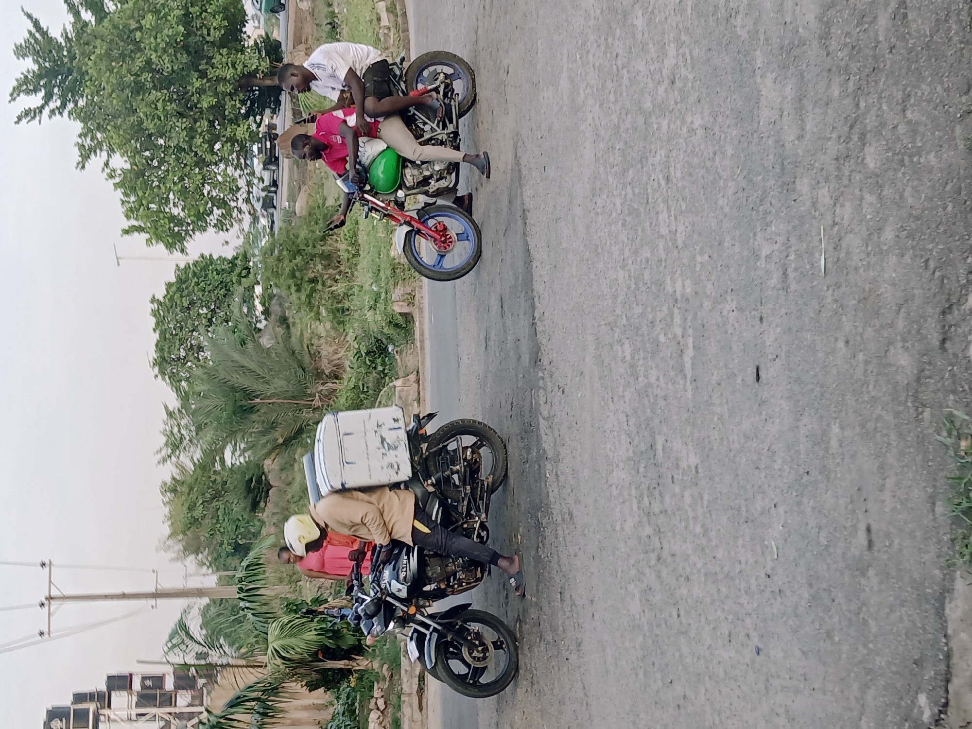 The scene depicts a road in Karum, Nigeria, with two motorcycles. The first motorcycle has two occupants, one wearing a white shirt and the other a red shirt. The motorcycle's frame is a green hue. The other motorcycle, carrying a white container, has two occupants, one wearing a tan shirt and the other a red shirt. The surrounding environment includes trees, plants, and a building.