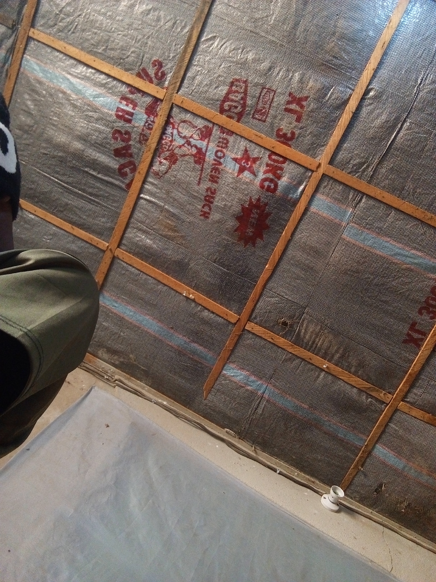 This image depicts an interior scene, likely a makeshift or unfinished structure. The dominant elements are a dark, woven material stretched taut and secured by wooden battens, forming a wall or ceiling. The material appears to be some kind of fabric or sheeting, possibly for insulation or covering, with red lettering and graphics visible in some areas. The wood battens are roughly cut and irregularly placed, suggesting a rudimentary construction method. In the lower left of the frame, a person's leg, clad in green trousers, is visible, indicating a human presence. The floor or ground below is covered by a light-colored, possibly tarpaulin-like sheet. A white light fixture is attached to a surface in the lower right portion of the image. The overall impression is one of a basic, possibly temporary, shelter. The context provided places this scene in Nyabe, Nigeria.