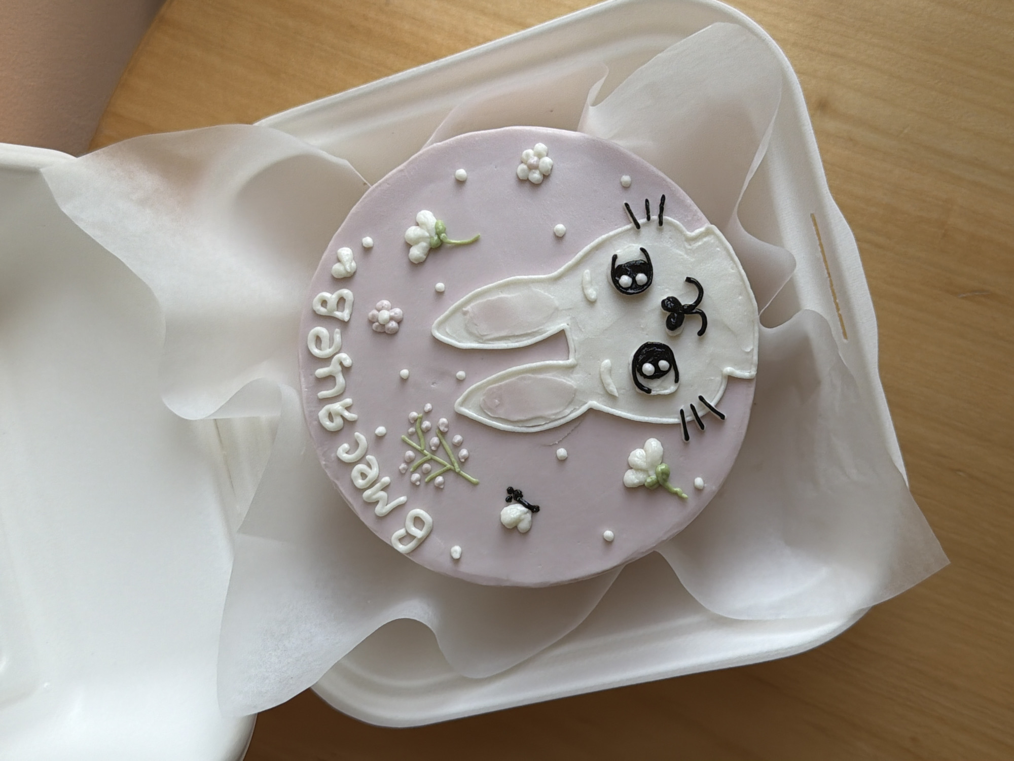 This image presents a small, round cake housed within a white disposable takeout container, lined with white tissue paper. The cake features a light lavender-purple base and is adorned with charming decorations.
The central design is a white stylized bunny face with prominent black-outlined eyes, a tiny black nose, a squiggly mouth, and delicate black whiskers. Short black lines also suggest eyebrows or eyelashes. Surrounding the bunny, the cake is decorated with various white and light pink icing flowers, some with green stems, and a delicate green sprig with white berry-like elements. Numerous tiny white icing dots are scattered across the purple surface, enhancing the whimsical theme.
On the left side of the cake, written in white icing using Cyrillic script, is the phrase "9 месяцев" (9 mesyatsev), which translates to "9 months." This indicates the cake is likely celebrating a baby's nine-month milestone, a common commemorative event.
The cake is placed on a light wooden surface, suggesting an indoor setting like a table or countertop. The lighting is soft and natural, evenly illuminating the cake and its intricate details. The overall aesthetic is one of cuteness and celebration, fitting for a child's special occasion. Given the context of Tel-Aviv, Israel, the Cyrillic text likely caters to the city's diverse population.
