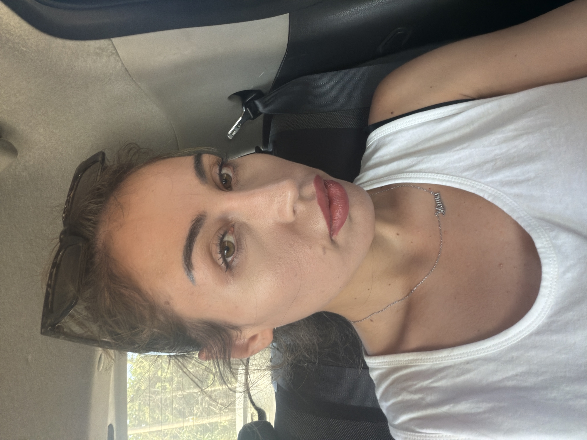 A woman is seated in the back of a vehicle, possibly a car, with a seatbelt fastened behind her. She is looking towards the camera, and her face is partially illuminated by natural light coming from an unseen window. Her hair is pulled back, and she is wearing sunglasses on top of her head. She has on a white tank top and a silver necklace with a nameplate. The background shows the interior of the car, including a headrest and upholstery, as well as a blurred view of trees and foliage through a window, suggesting an outdoor environment. The scene appears to be located in Dromolaxia - Meneou Municipality, Cyprus, given the context provided.
